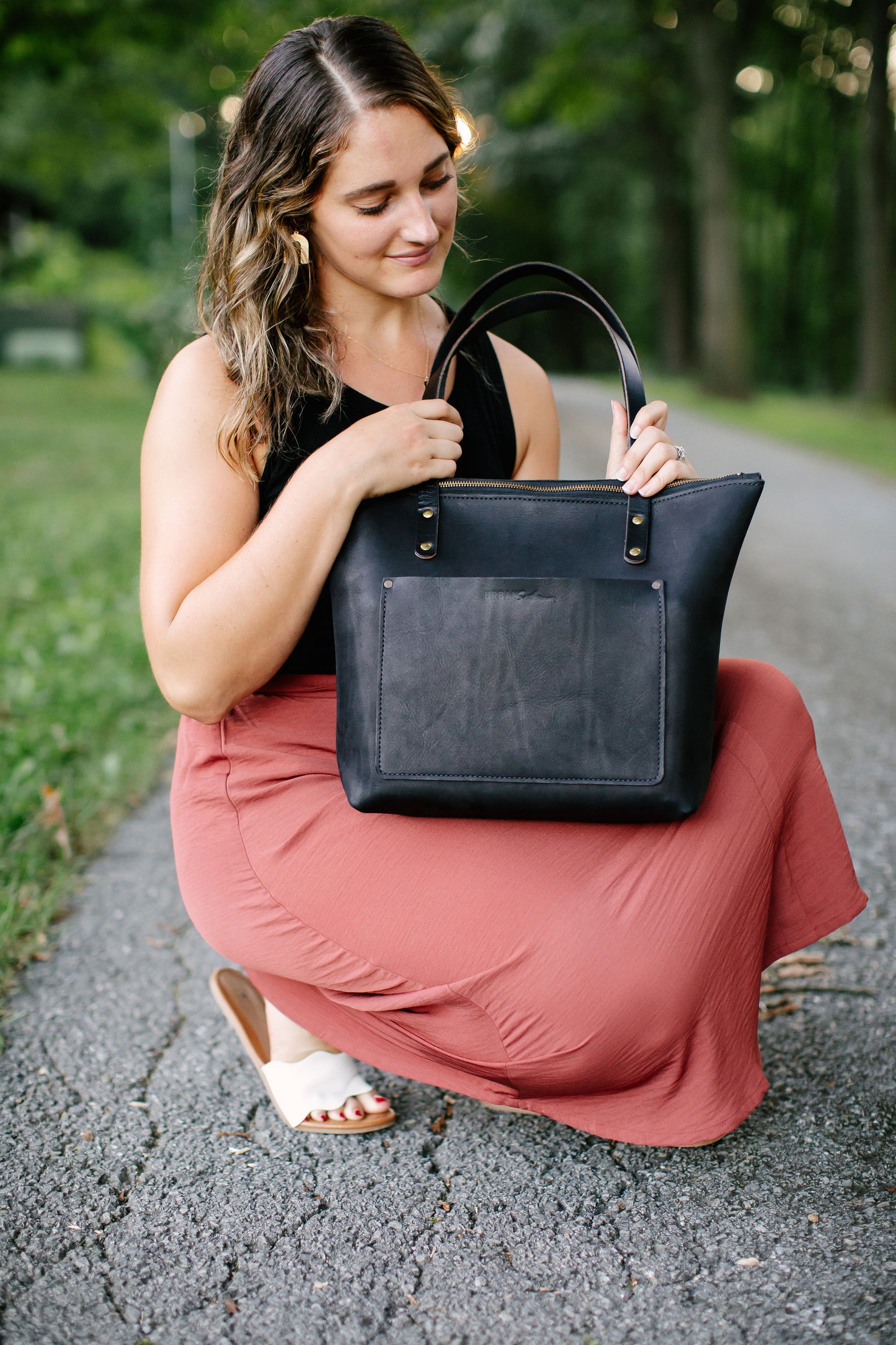 A woman kneels down, resting her black Urban Zipper Tote on her knee. She begins opening the leather bag.