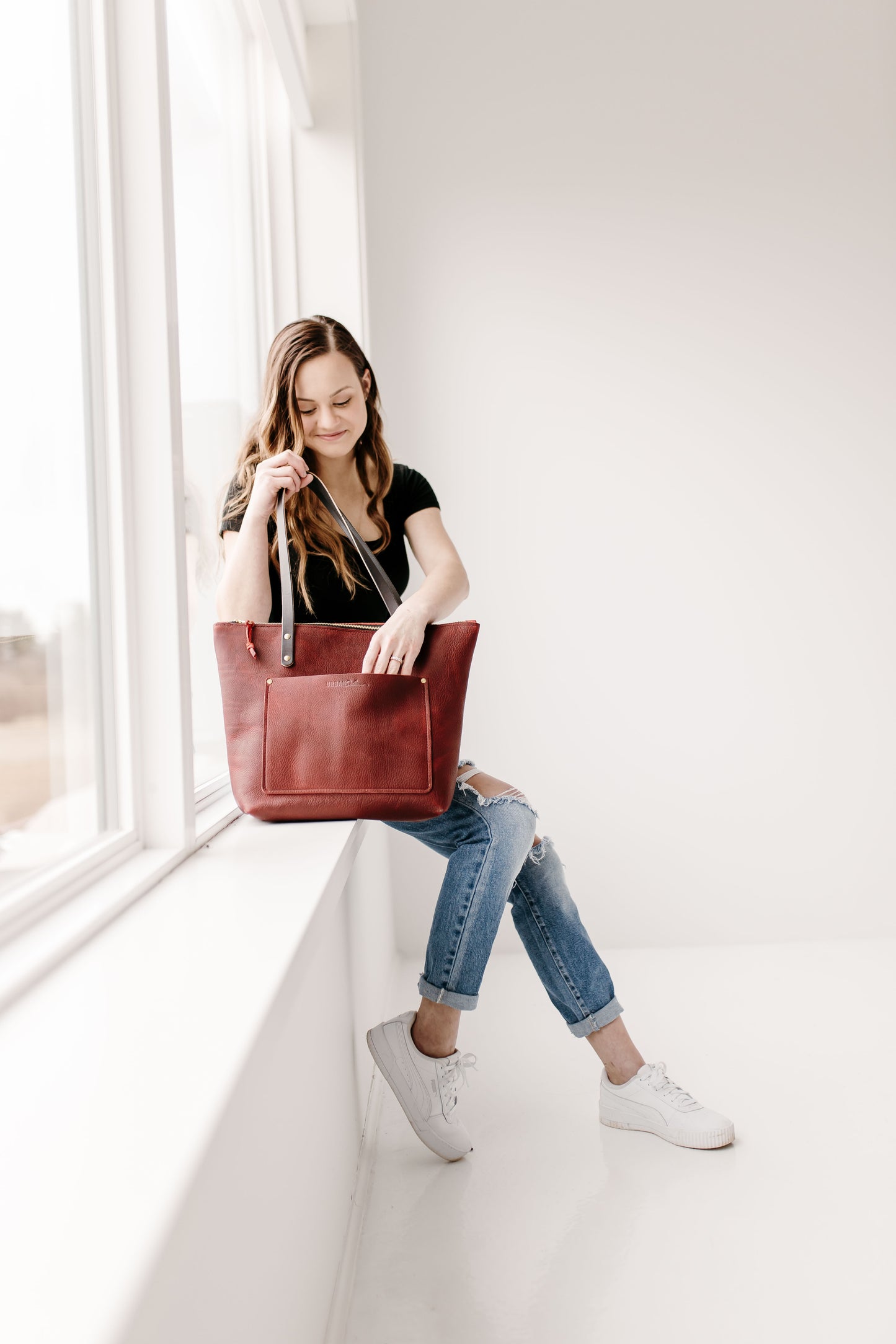 A woman sitting on a windowsill, holds a merlot Urban Zipper Tote by her side, while reaching into it's large front slip pocket.
