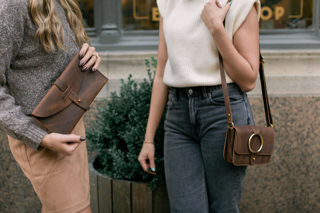 A woman holds the 5th Avenue Clutch in Rustic Brown while standing next to another woman carrying the 5-Pocket Crossbody bag.