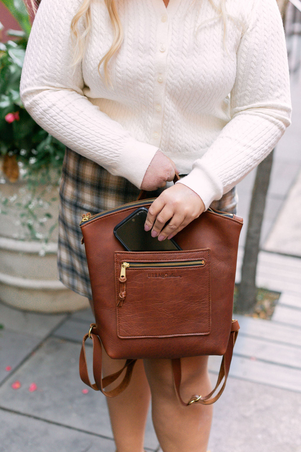 A woman slips her smartphone into the front exterior slip pocket on the Brooklyn Backpack in Cognac.