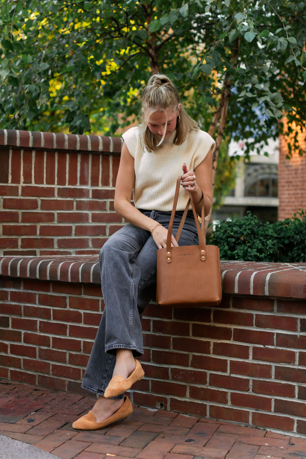 A woman sitting on a wall reaches her hand into her Honey Mini Urban Tote. 
