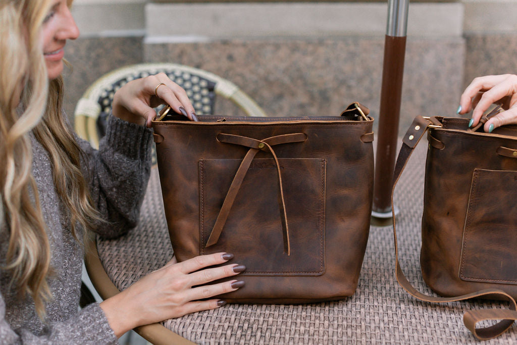 A woman sitting at a table begins unzipping her rustic brown Lexington Bow handbag. 