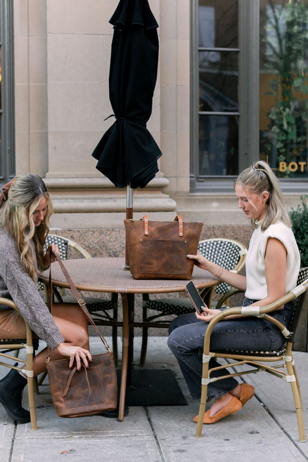 Two women sit at an outdoor cafe. One woman sets her rustic brown Lexington Bow handbag on the ground, while the other woman looks at her phone, her rustic brown Urban Zipper tote sits on the table next to her. 