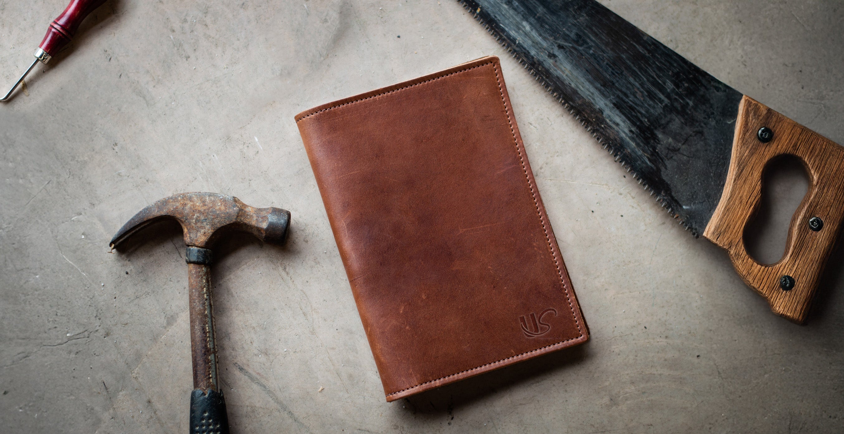 A leather notebook lays on the ground next to a hammer & saw. 