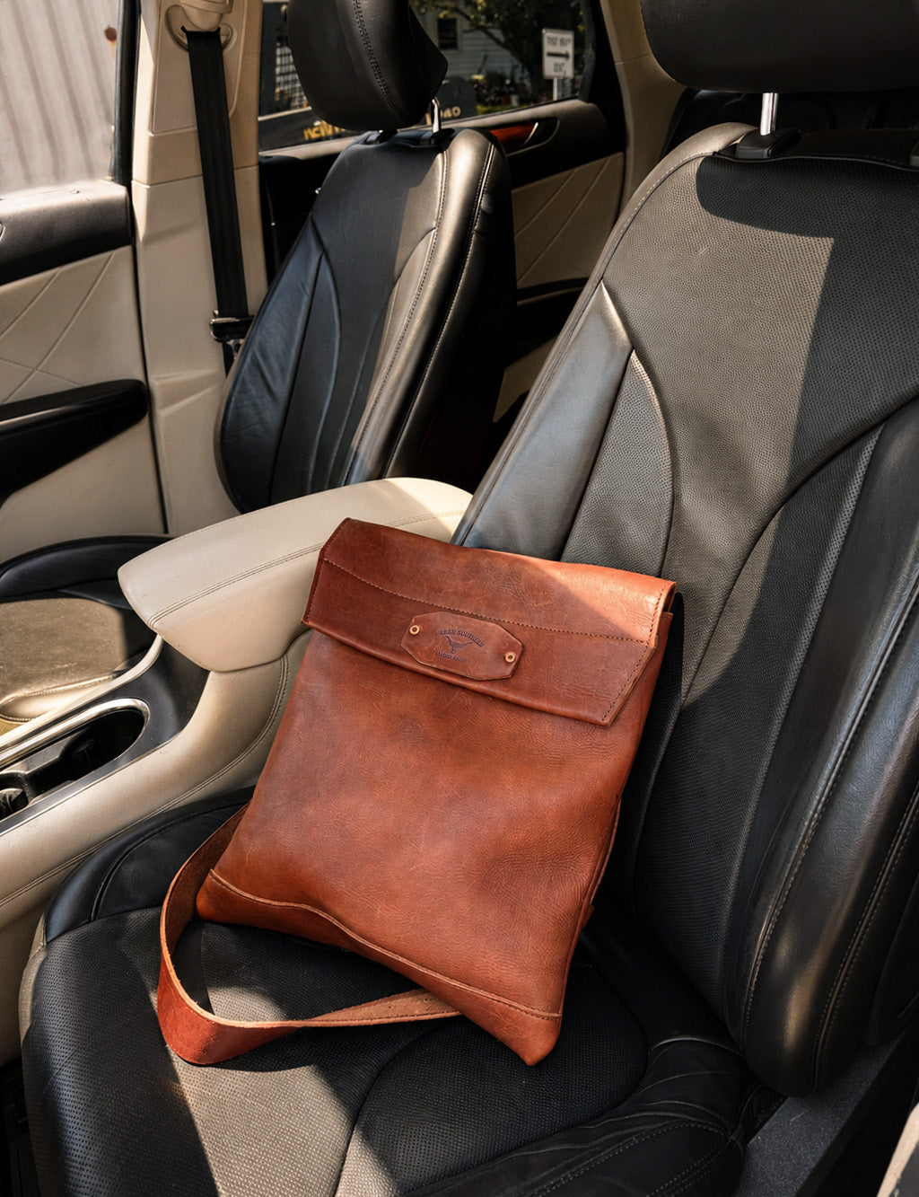 A brown leather shoulder bag with a flap closure resting on a black and gray car seat.
