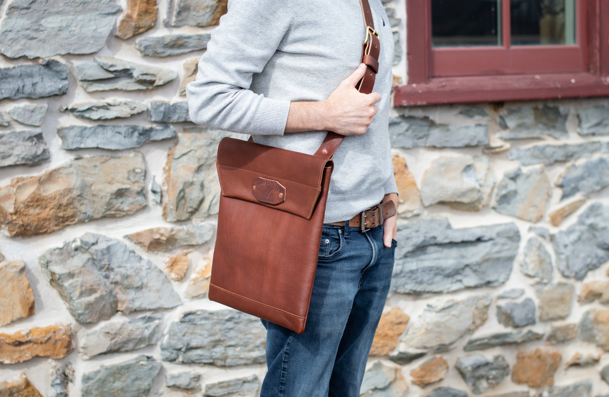 Man holding a brown leather courier bag on his shoulder against a stone wall.