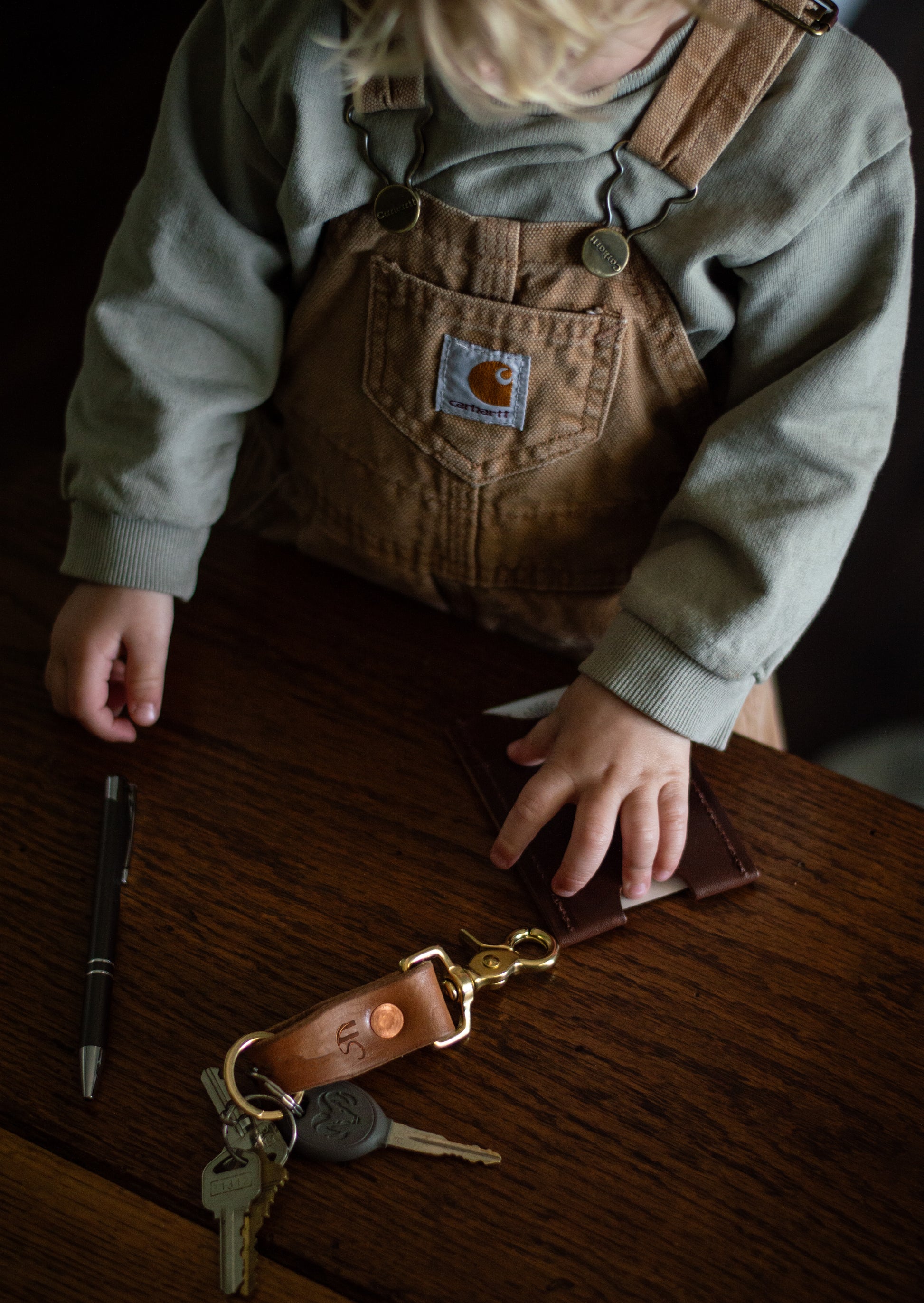 A child wearing brown Carhartt overalls and a gray sweatshirt sits at a dark wooden table, holding a brown leather keychain with gold-tone hardware. 