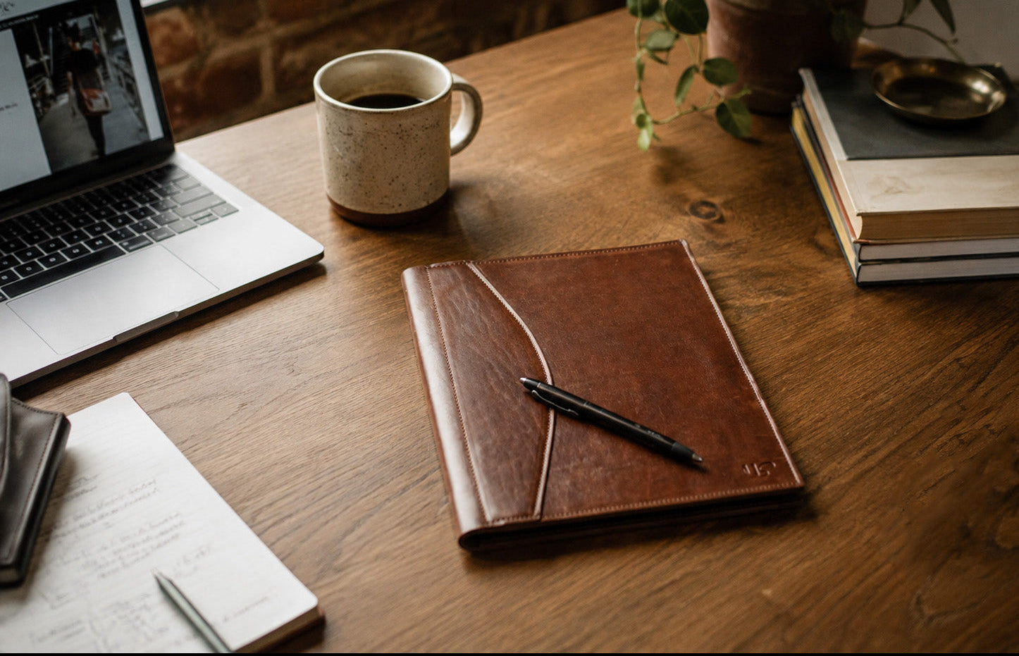 A brown leather padfolio with a pen resting on it, placed on a wooden desk next to a mug of coffee, an open laptop, books, and a potted plant.