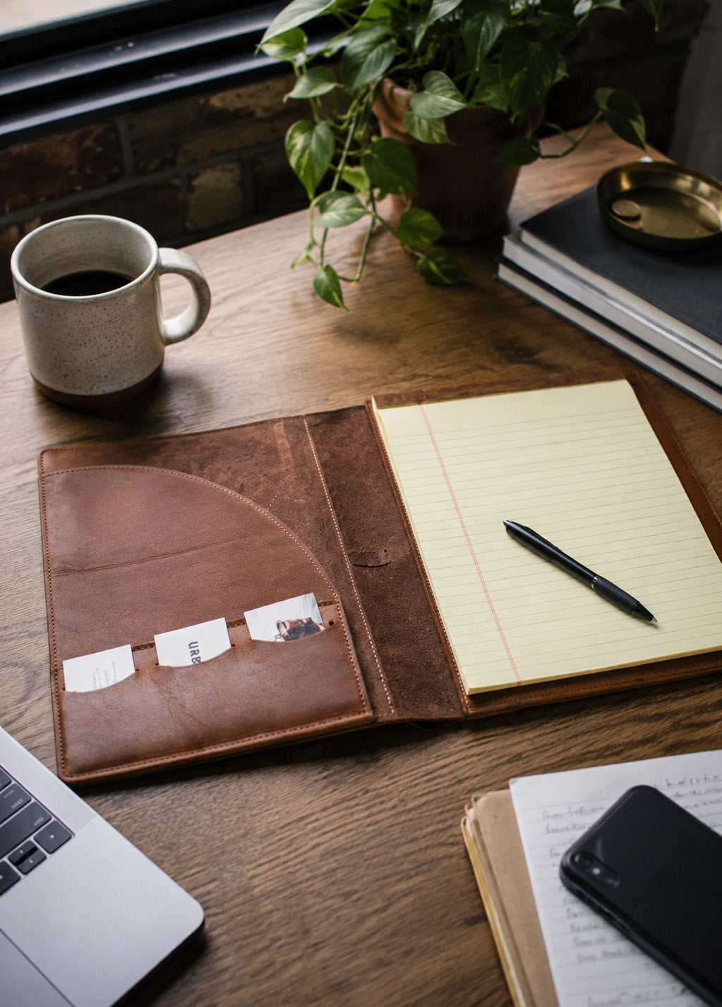 A brown leather padfolio laid open on a wooden desk, holding a lined notepad and a black pen. A mug of coffee, a potted plant, books, and part of a laptop are visible around it in a styled workspace scene.