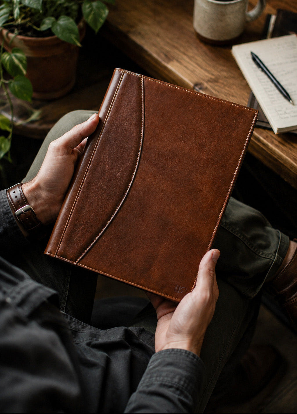 A person holds a brown leather padfolio with a curved seam detail, shown in a cozy indoor setting with a wooden table, mug, pen, and plant in the background.