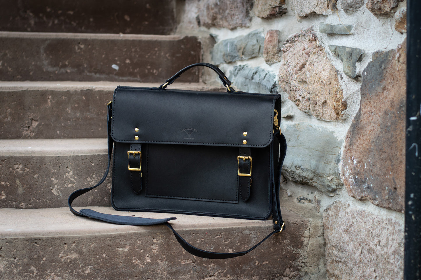 A black leather Executive Briefcase sits on a step, it's sturdy & comfortable shoulder strap lays beside it. 