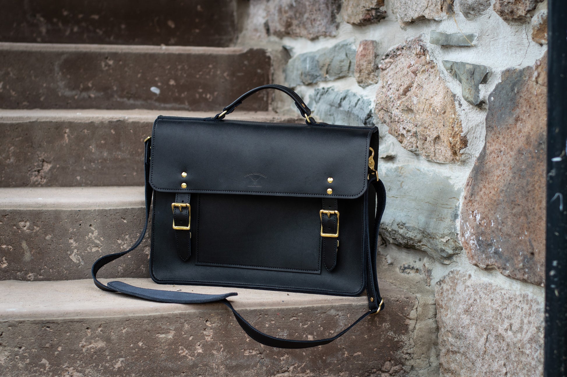 A black leather Executive Briefcase sits on a step, it's sturdy & comfortable shoulder strap lays beside it. 
