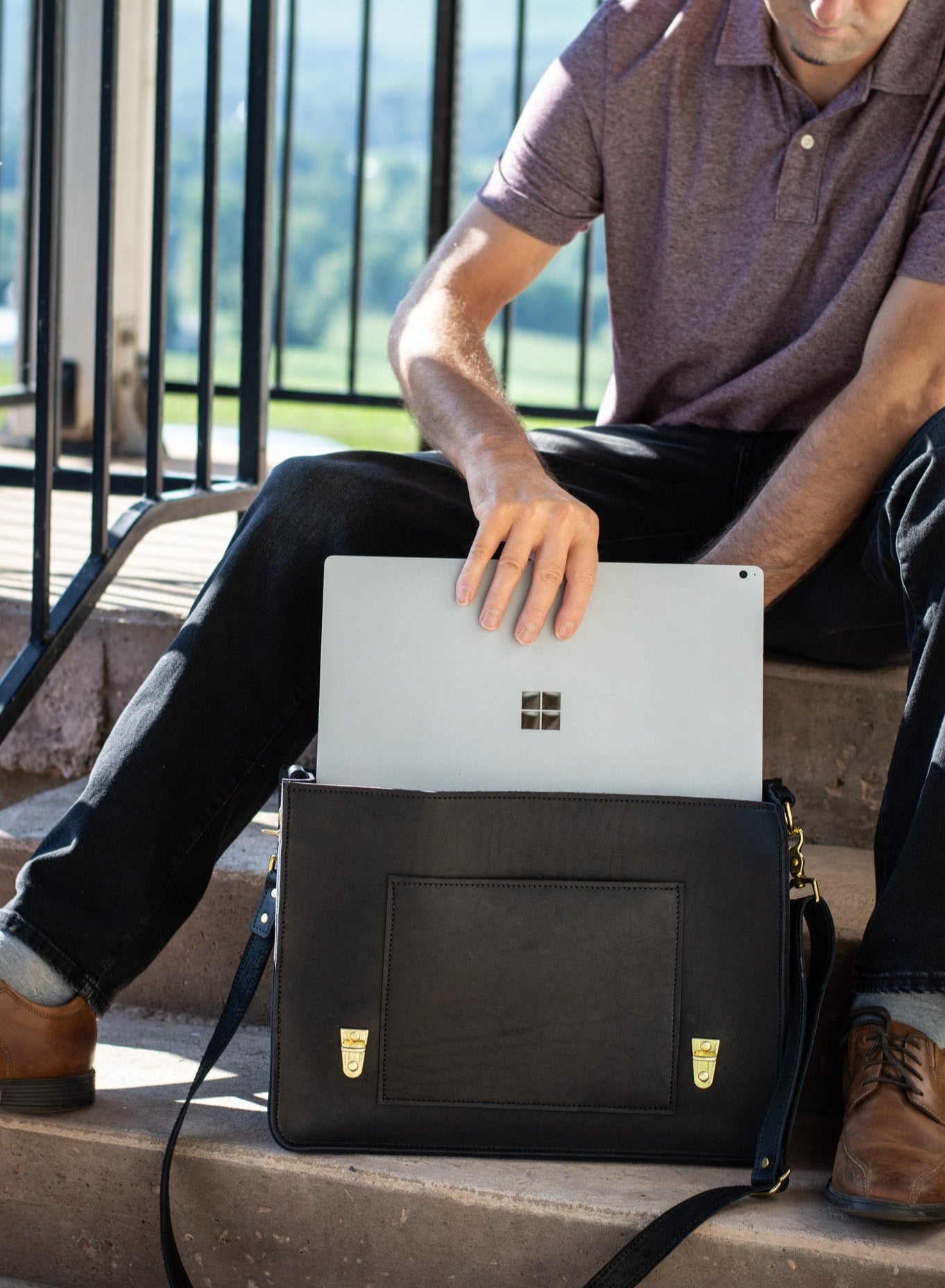 A man sits on a step with a black Executive Briefcase by his side. He reaches into the spacious interior of the satchel, pulling out his laptop. 