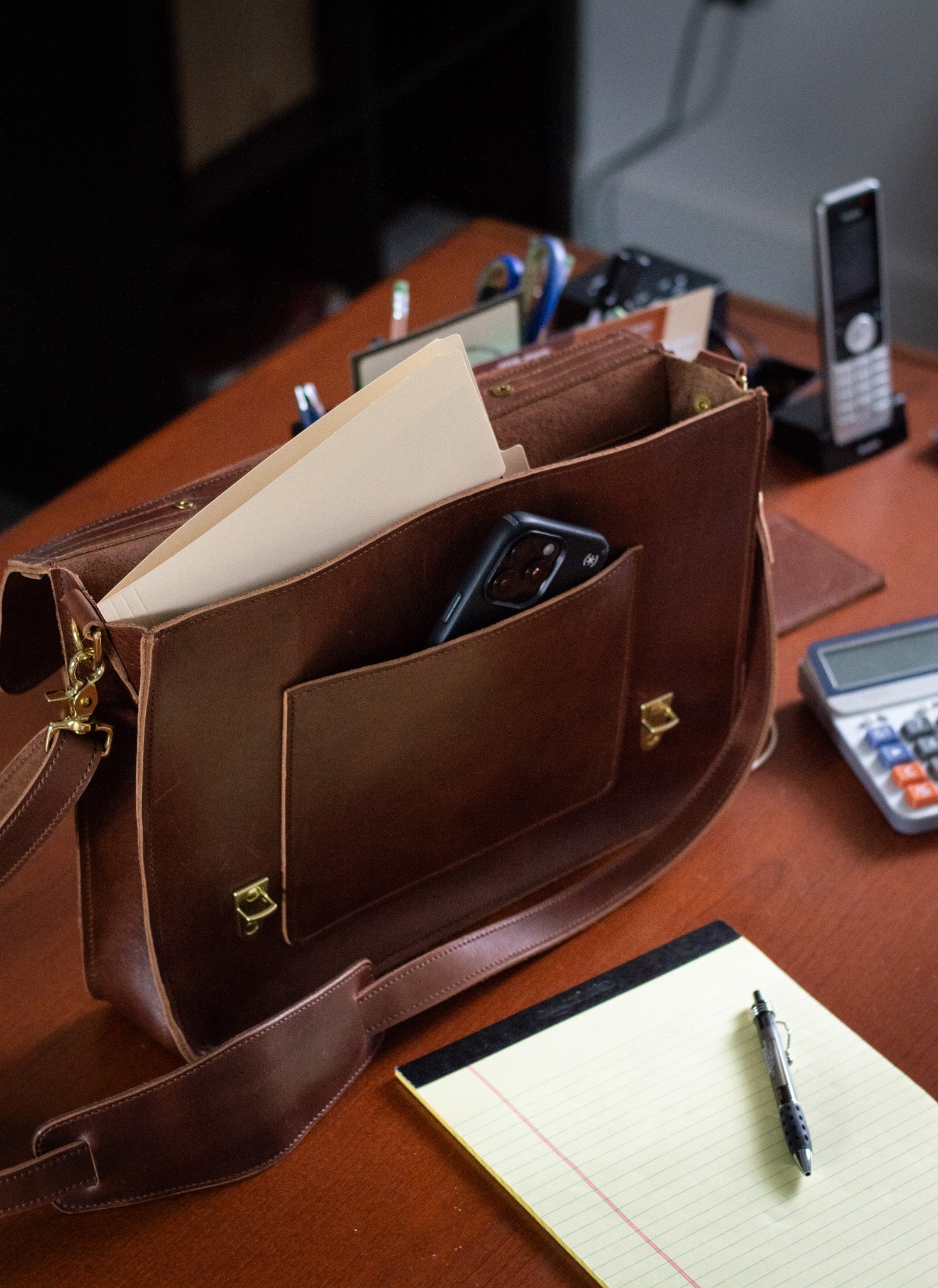 A Whiskey leather Executive Briefcase sits open on an office desk. An iphone is nestled in the exterior front pocket, while a folder peeks out of the interior of this spacious satchel. 