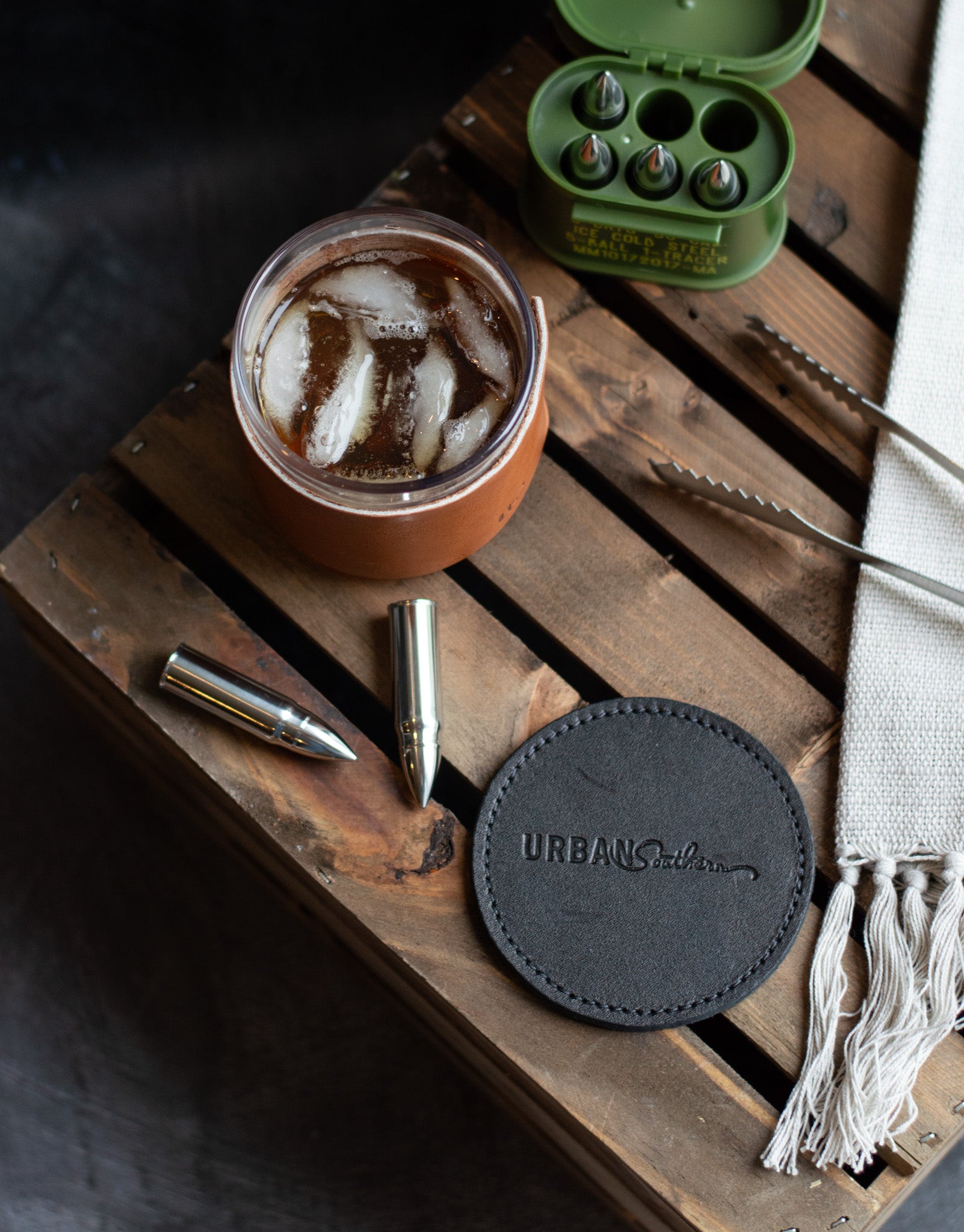 A Black, Urban Southern branded coaster sits on top of a wooden crate with a glass of whiskey next to it. 