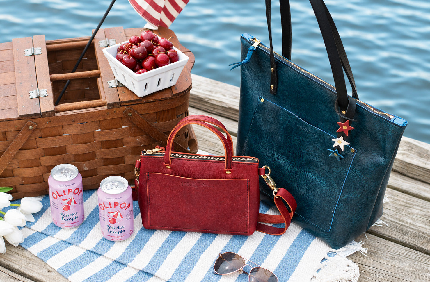 Two handbags, a picnic basket with cherries, and Olpico drinks on a wooden deck by water. A heritage star bag charm is looped around the handle of the blue zipper tote. 