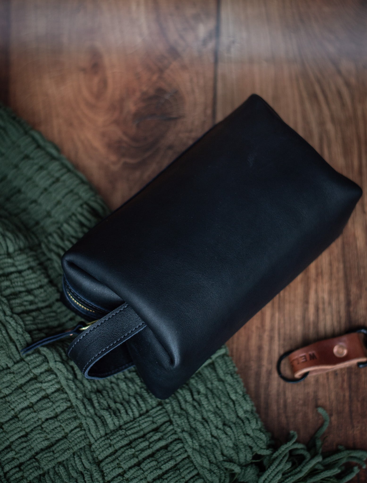 Black leather dopp kit laying on a hardwood floor with a green blanket next to it. 