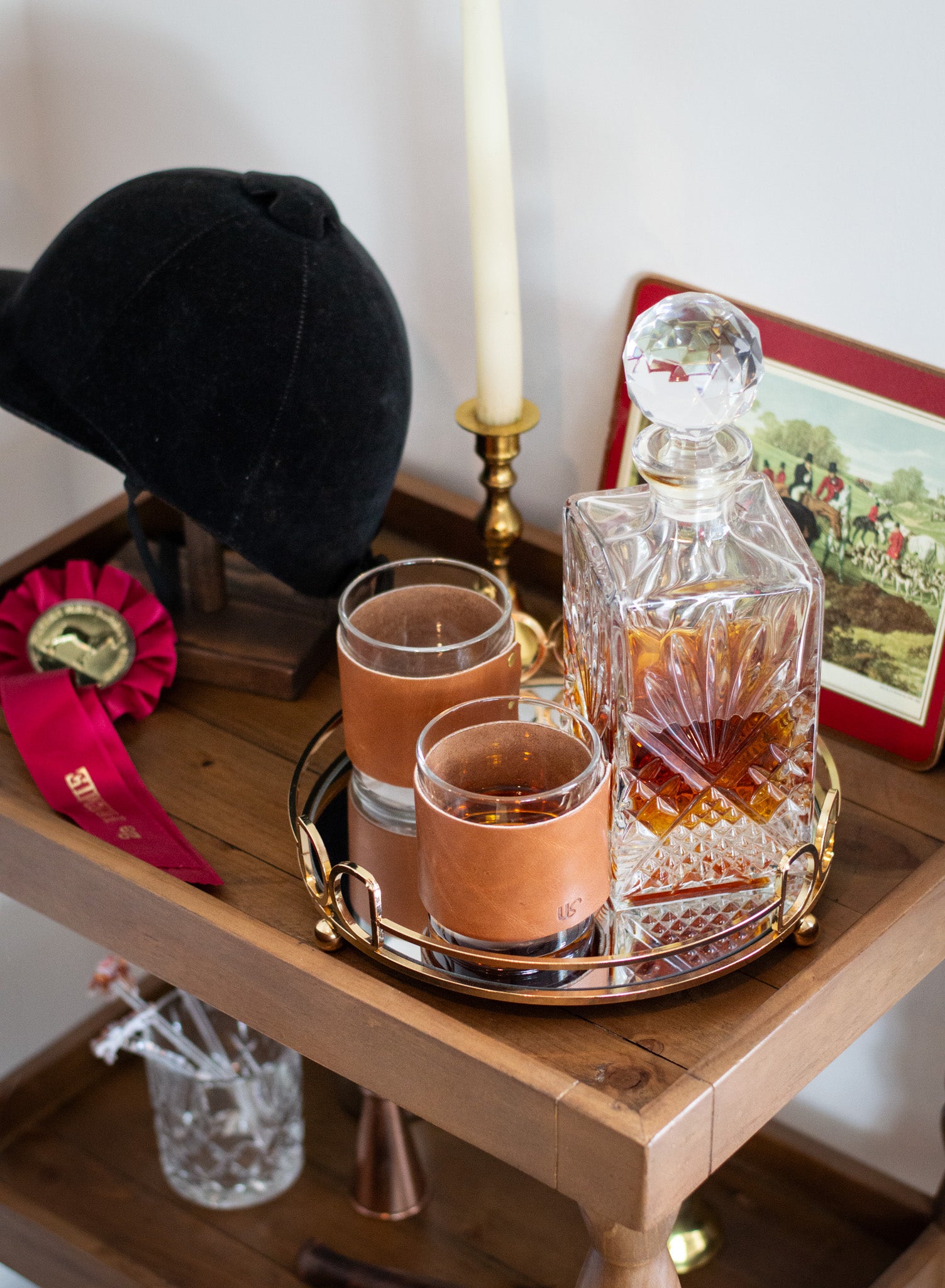 Two glasses wrapped in leather sleeves on a tray with a crystal decanter, candle, and decorative items on a wooden bar cart.