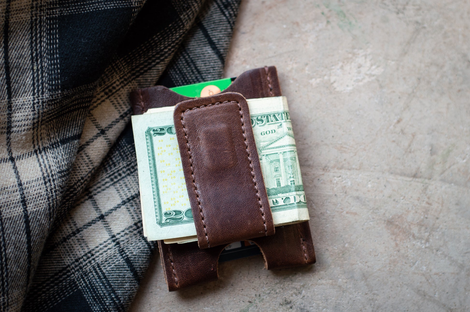 Brown leather money clip holding dollar bills on a textured surface with plaid fabric.