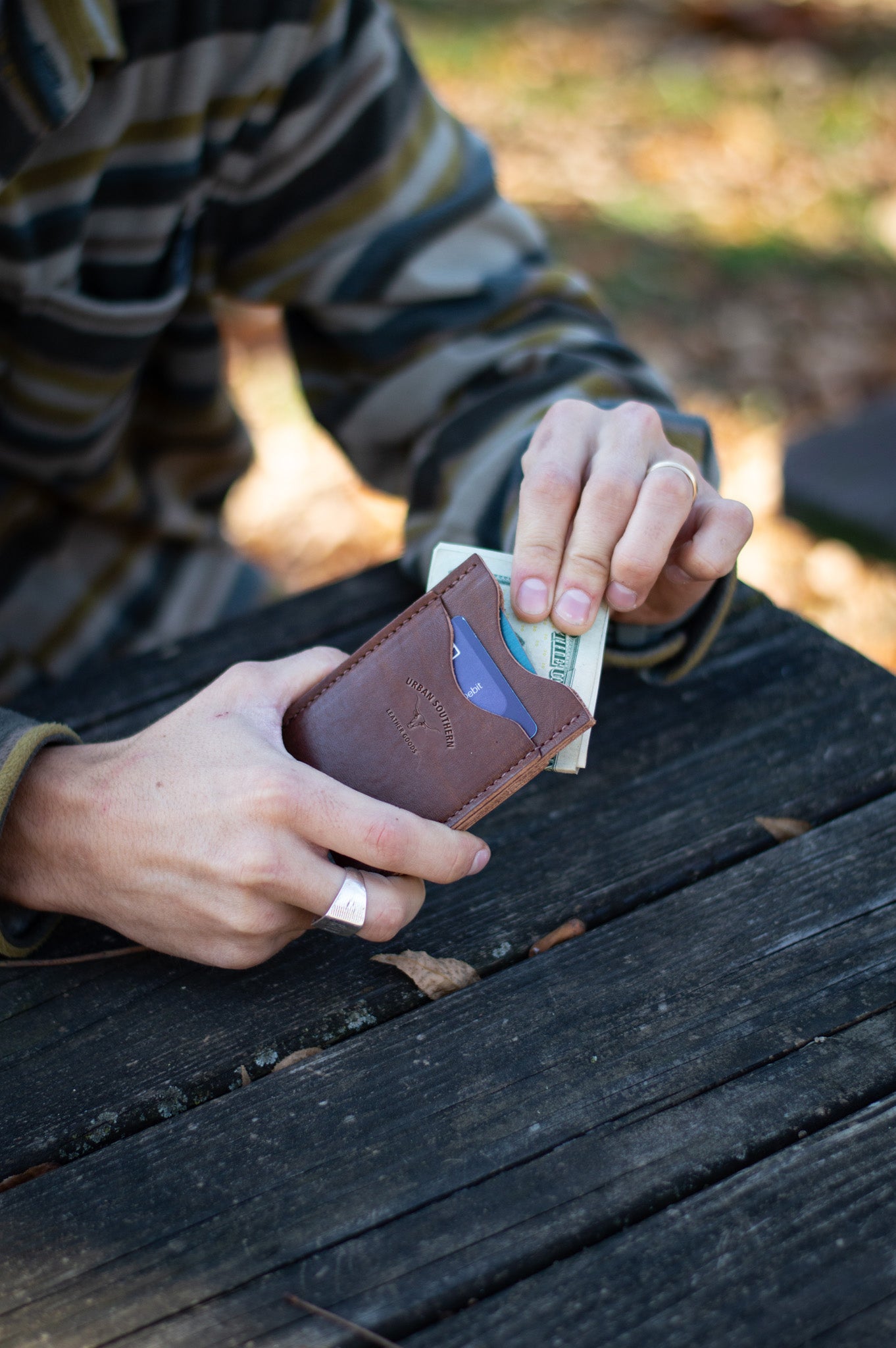 A man is holding a leather wallet, removing money from the magnetic clip, on a wooden surface outdoors