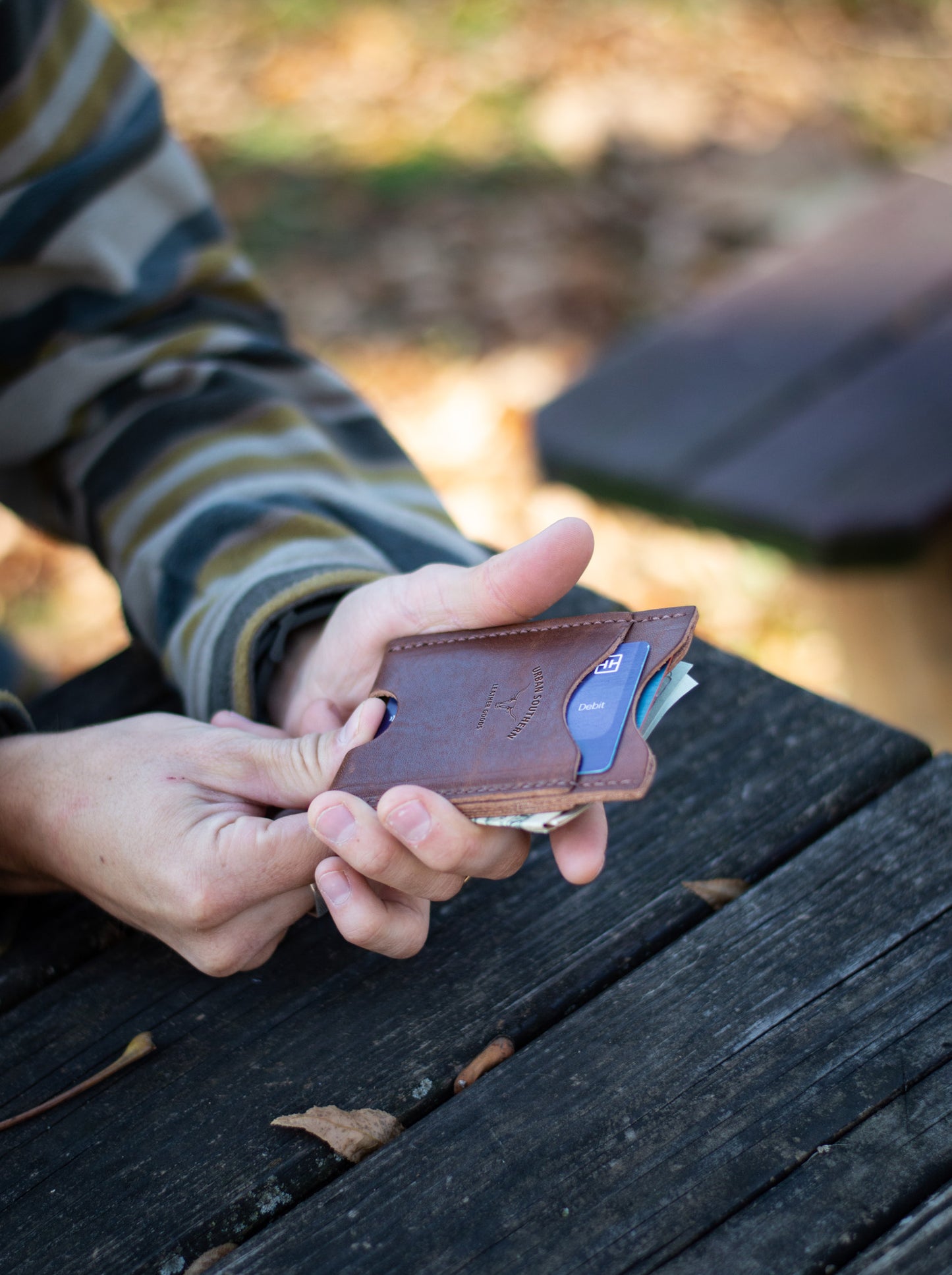 A man is holding a leather wallet, pushing out a credit card using the convenient bottom access notch, on a wooden surface outdoors