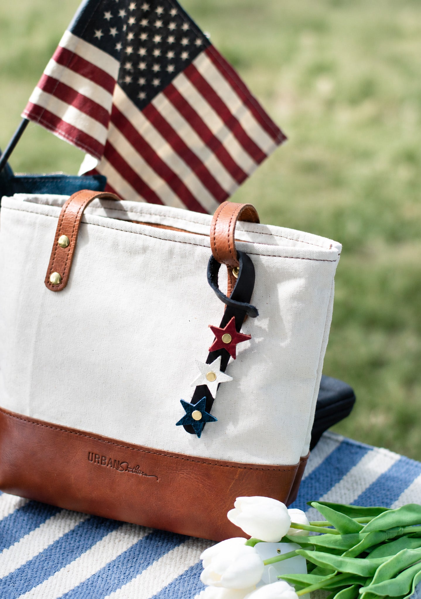 White tote bag with brown leather accents on a striped blanket with tulips and an American flag in the background. The heritage star bag charm is looped around the handle of the tote. 
