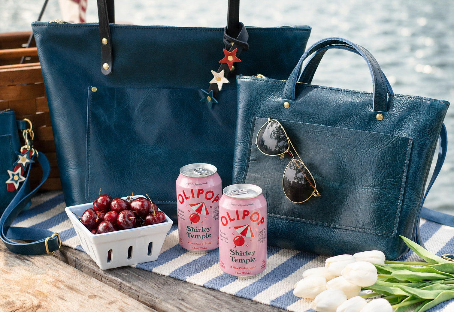 Two blue handbags, including the quinn crossbody with Olipos drinks and snacks on a table by water.