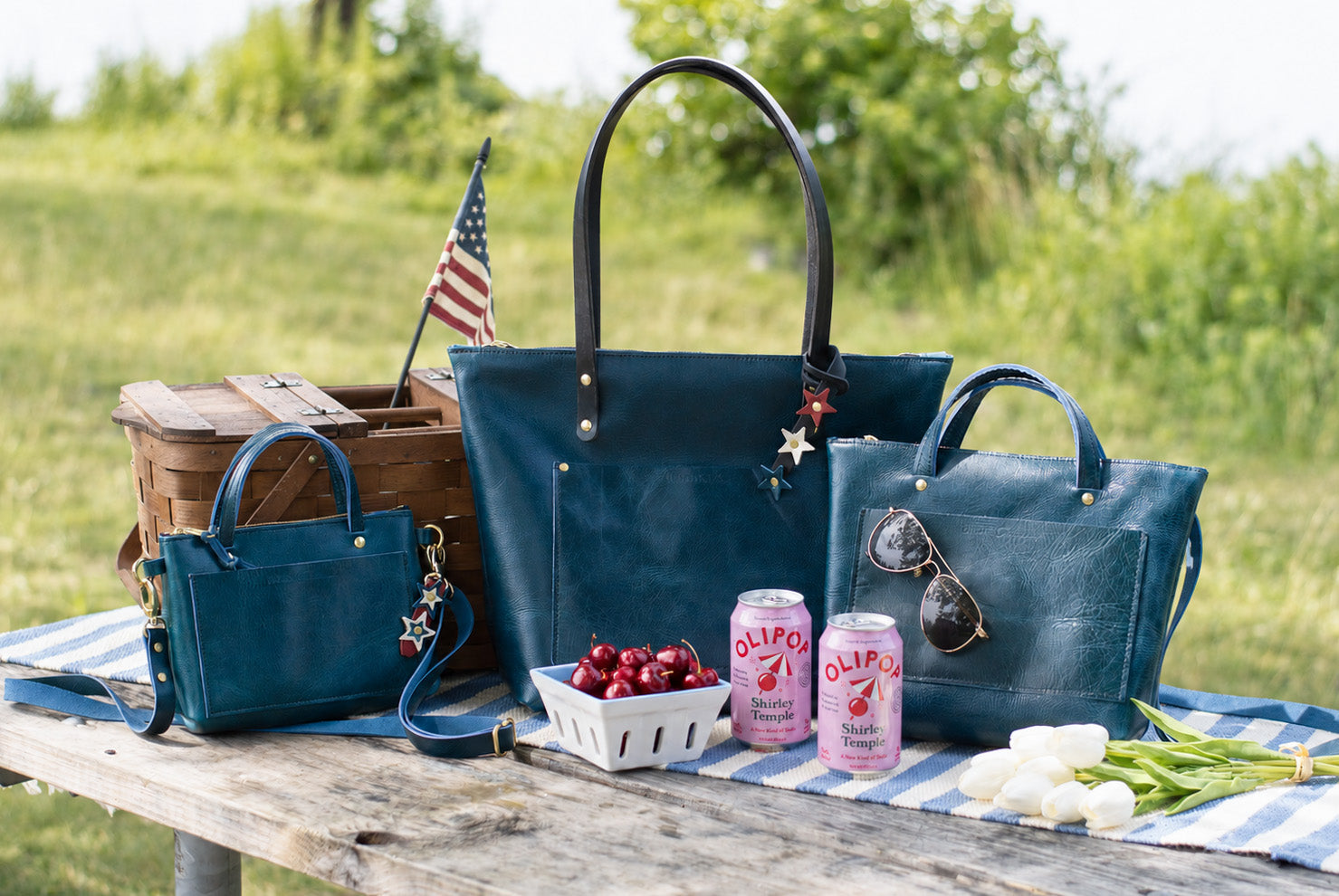 A selection of blue leather handbags, including the quinn crossbody, sit on a picnic table with picnic basket & food.