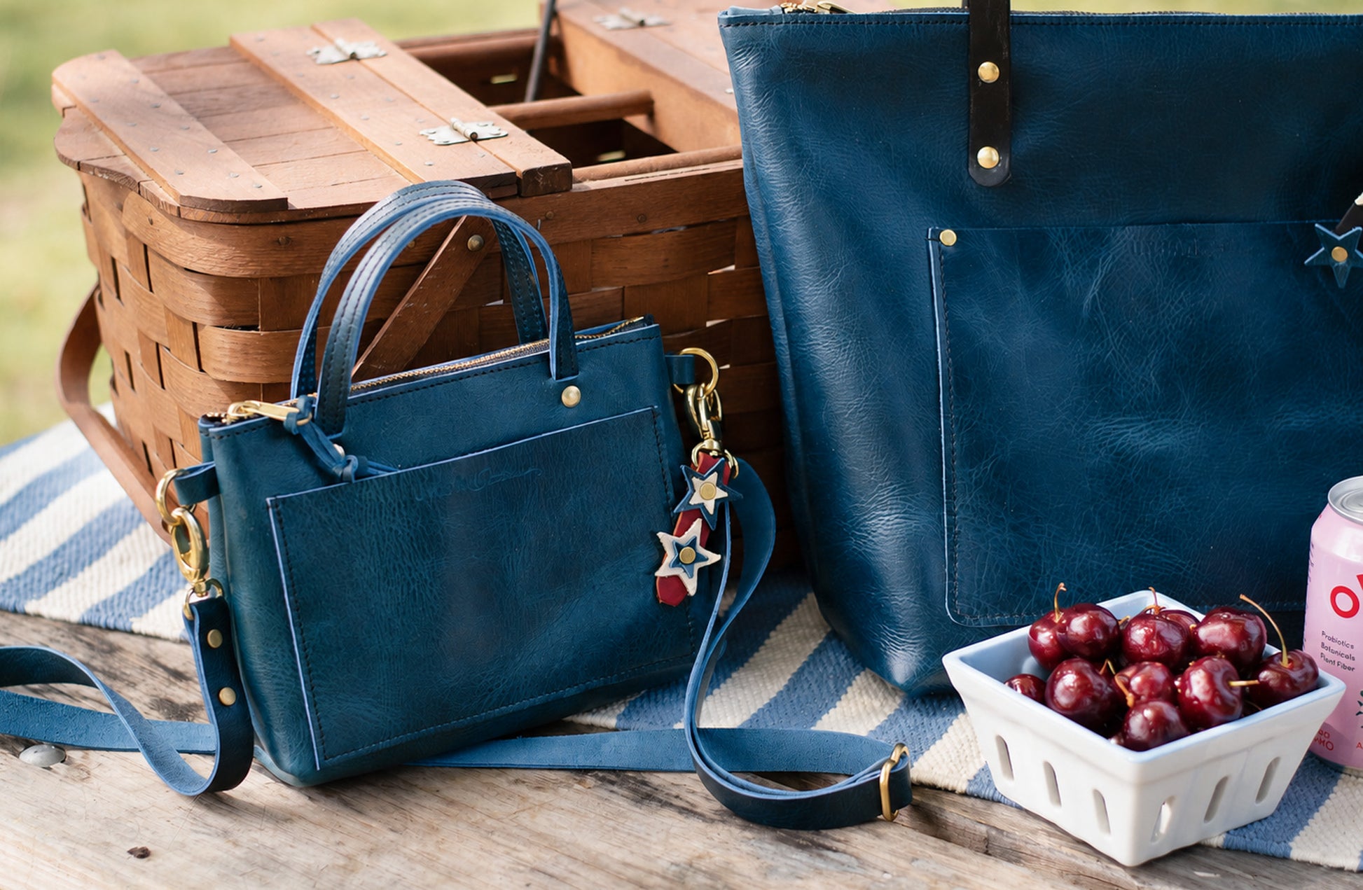 Two blue handbags on a picnic blanket with a basket and bowl of cherries in the background. A Jubilee bag charm is attached to a brass ring on the smaller handbag. 