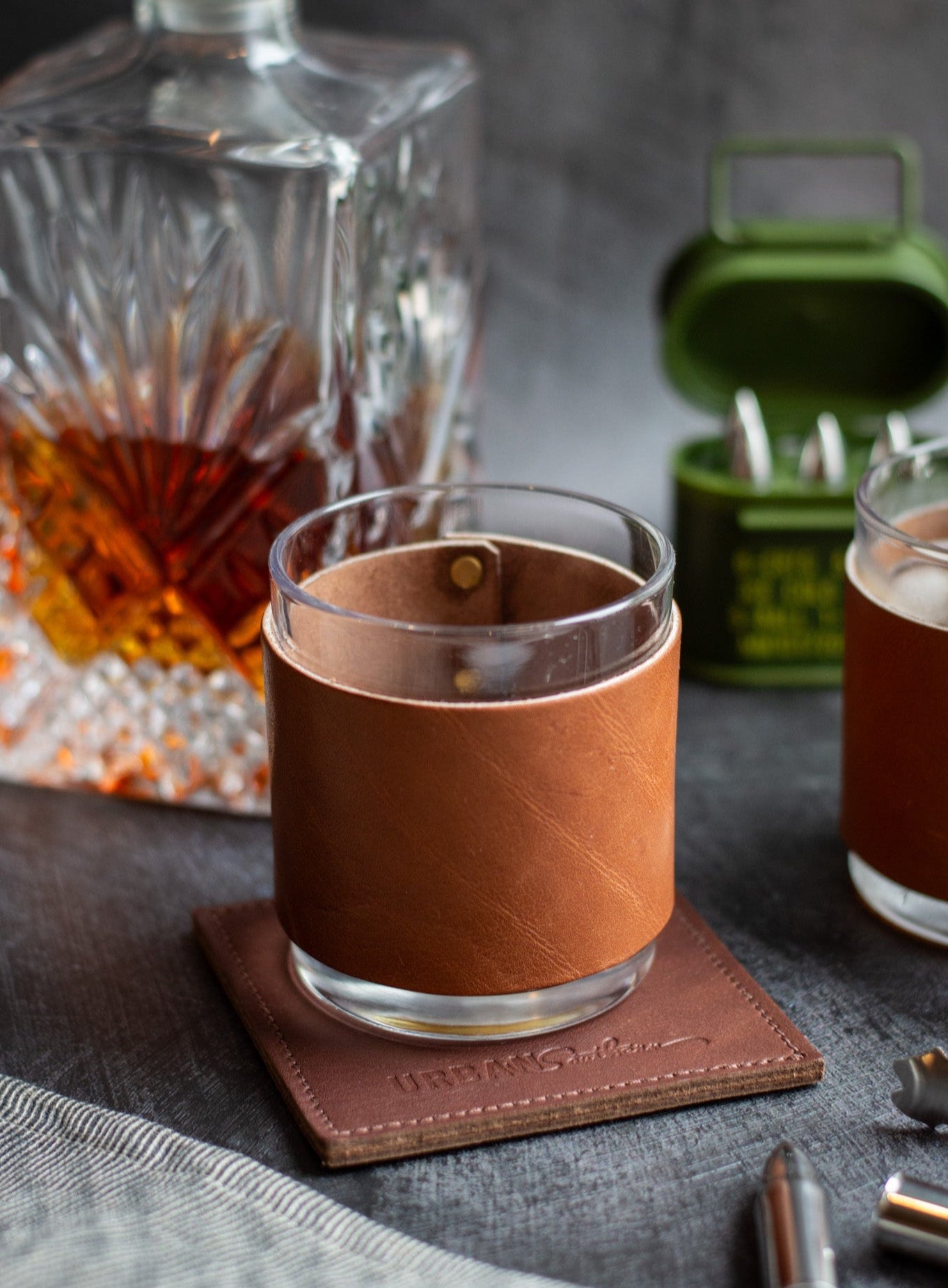 Glass with brown leather sleeve on a coaster, next to a decanter and another glass, on a textured surface.