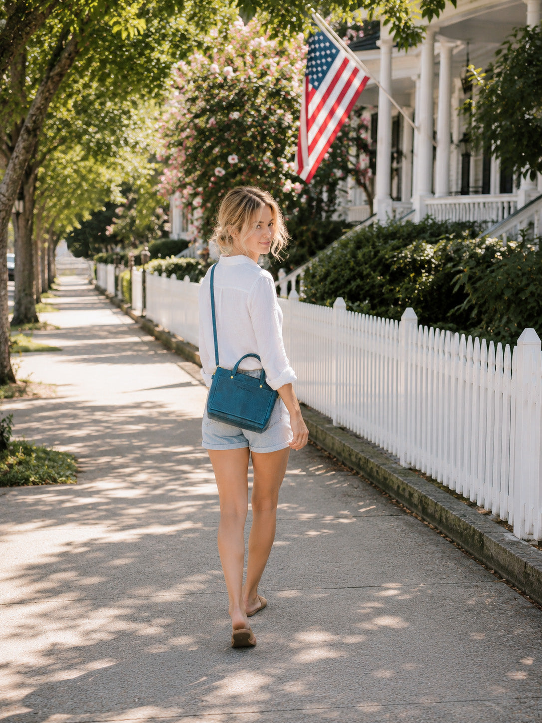 Woman walking on a sidewalk with a white picket fence and American flag in the background, wears a blue leather cassidy crossbody on her shoulder. 