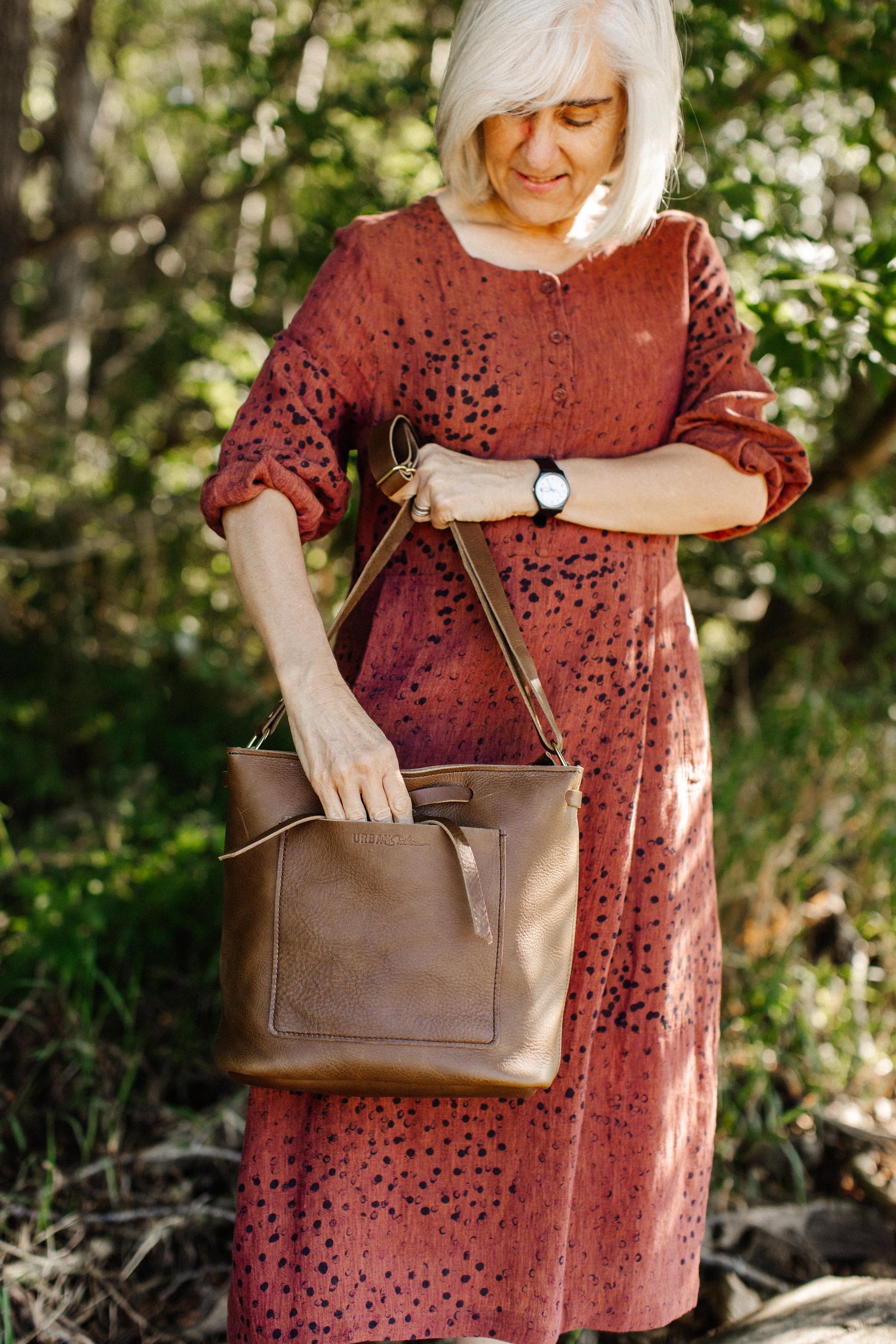 A woman holds a Lexington Bow leather handbag by its shoulder strap, while reaching into the spacious exterior front pocket. 