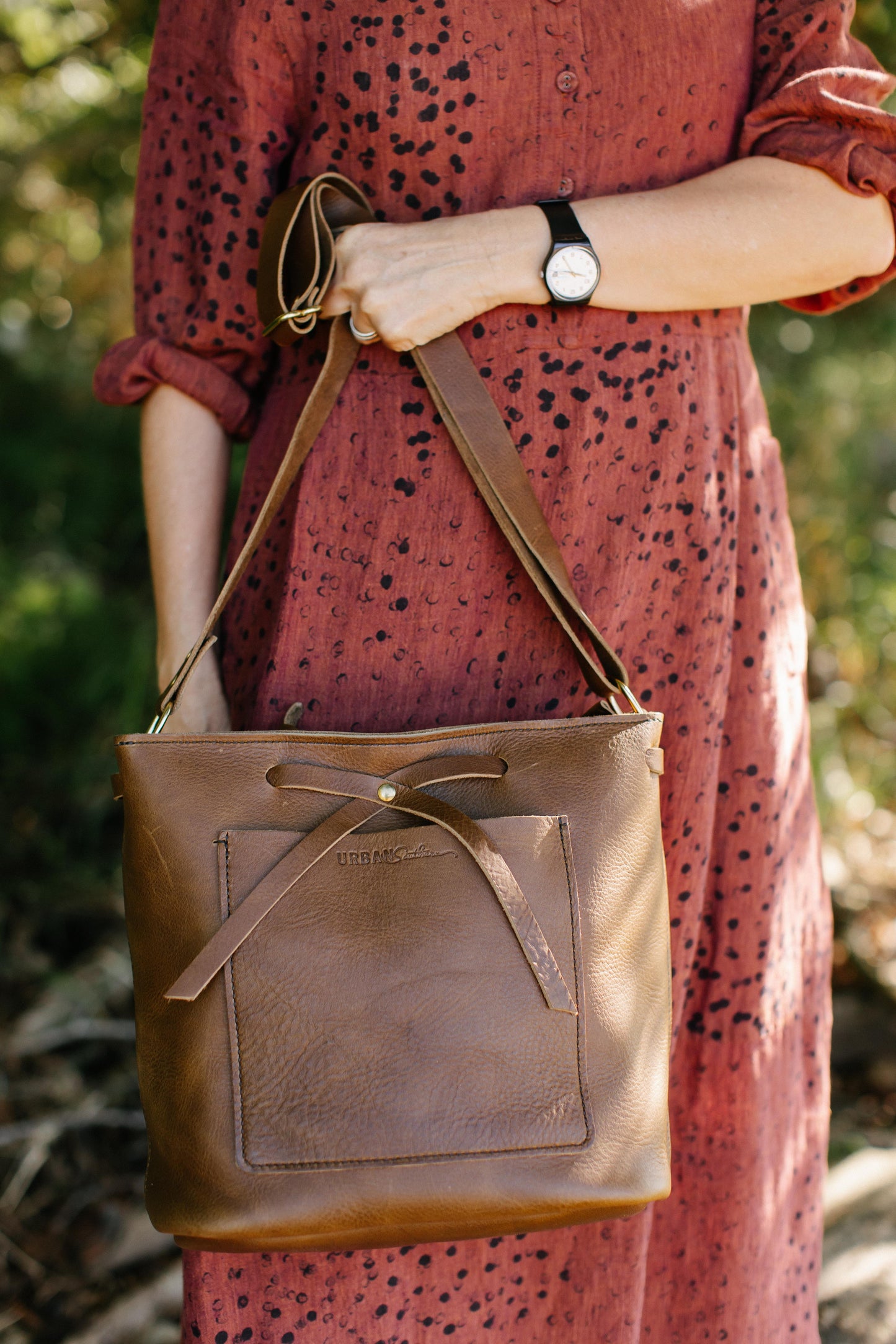 A woman holds a Lexington Bow handbag by its shoulder strap, showcasing the signature leather bow on the front of the bag. 
