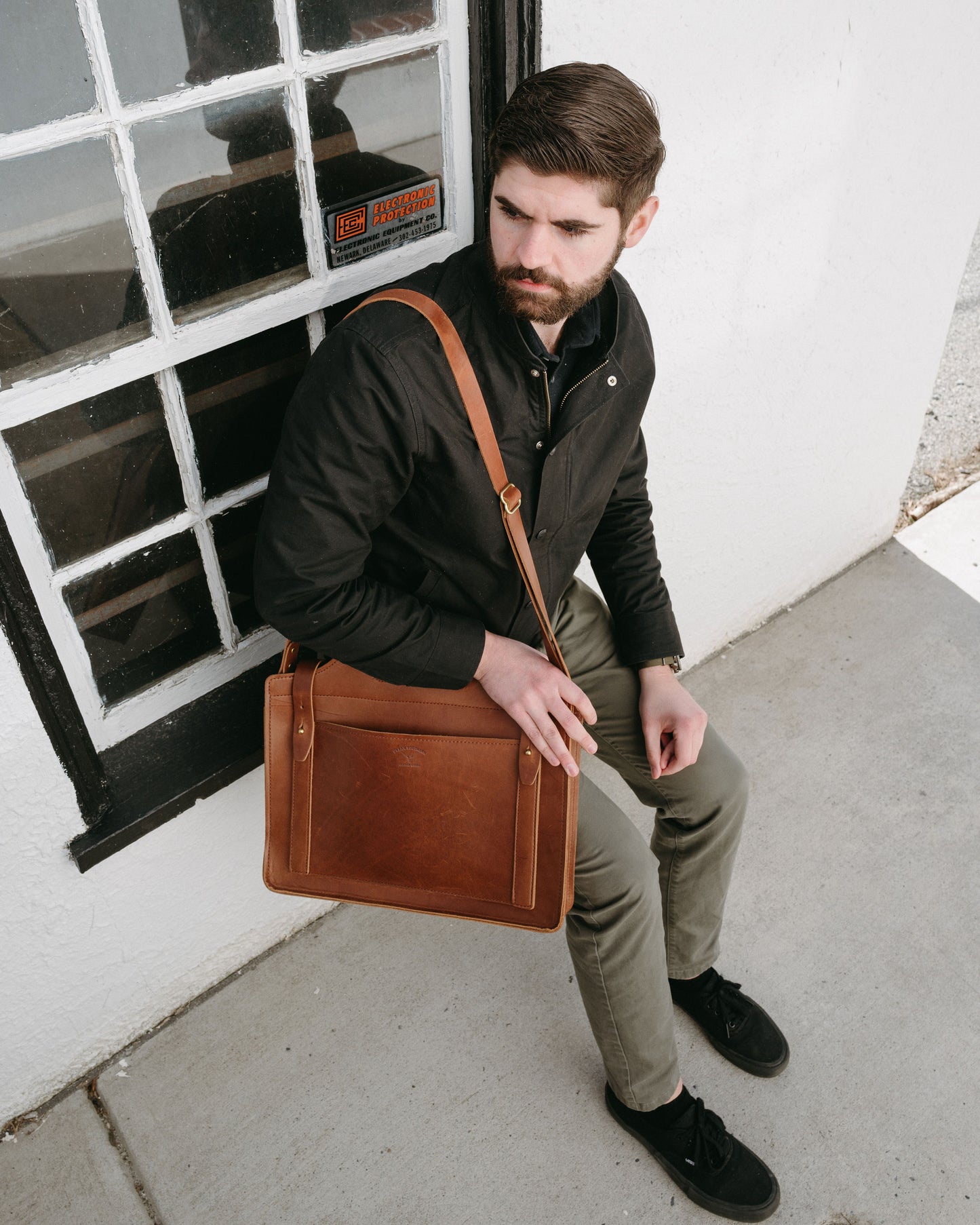 Man holding a brown leather satchel against a white wall with a window.