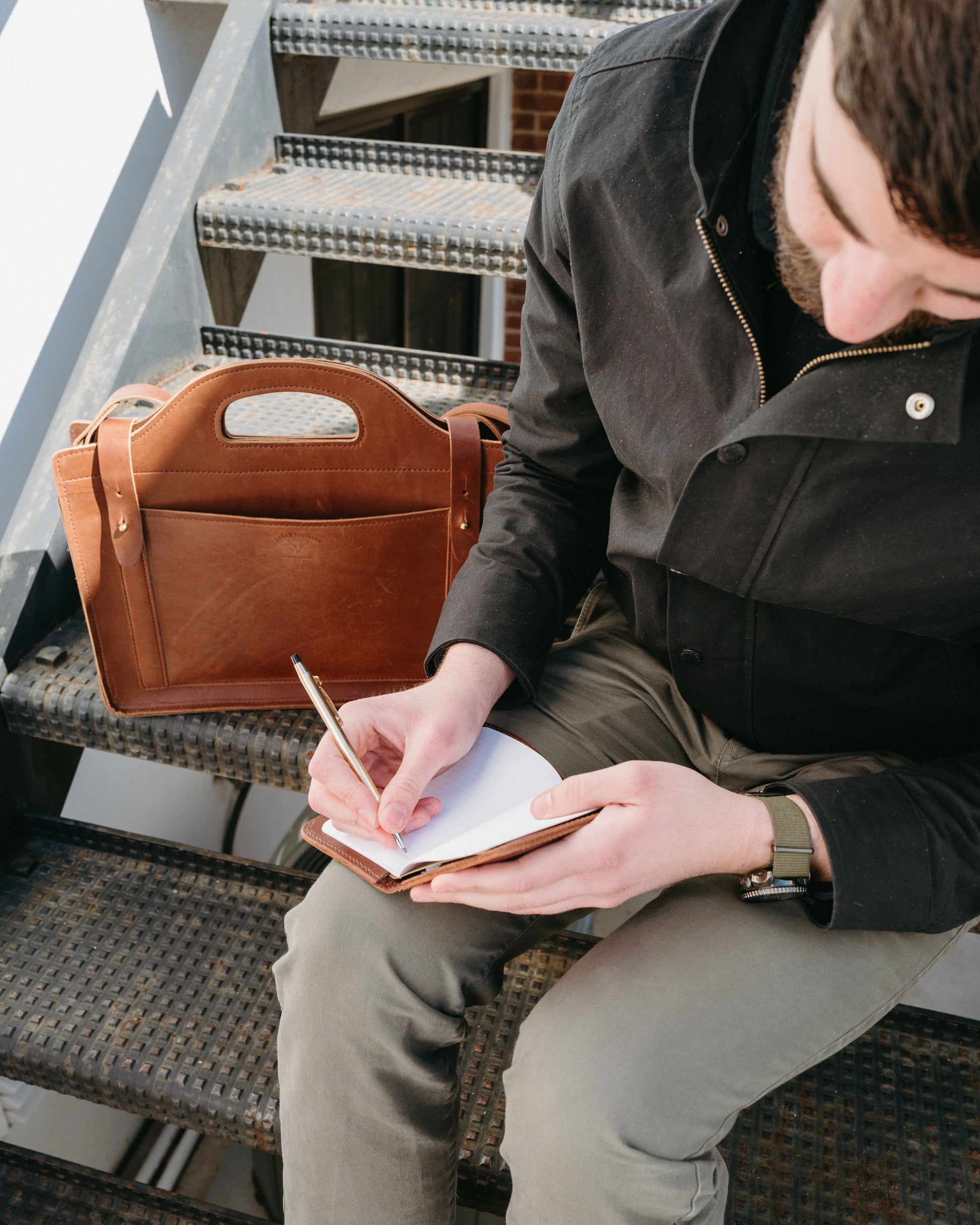 Person sitting on metal stairs writing in a notebook with a brown leather satchel beside them.