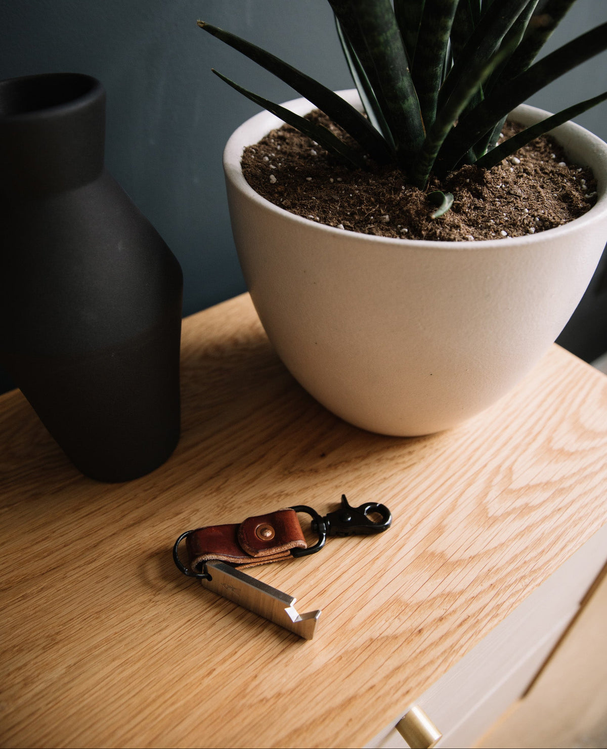 A leather keychain with a silver metal tool attached, resting on a light wood table next to a white planter with a green plant and a black vase.