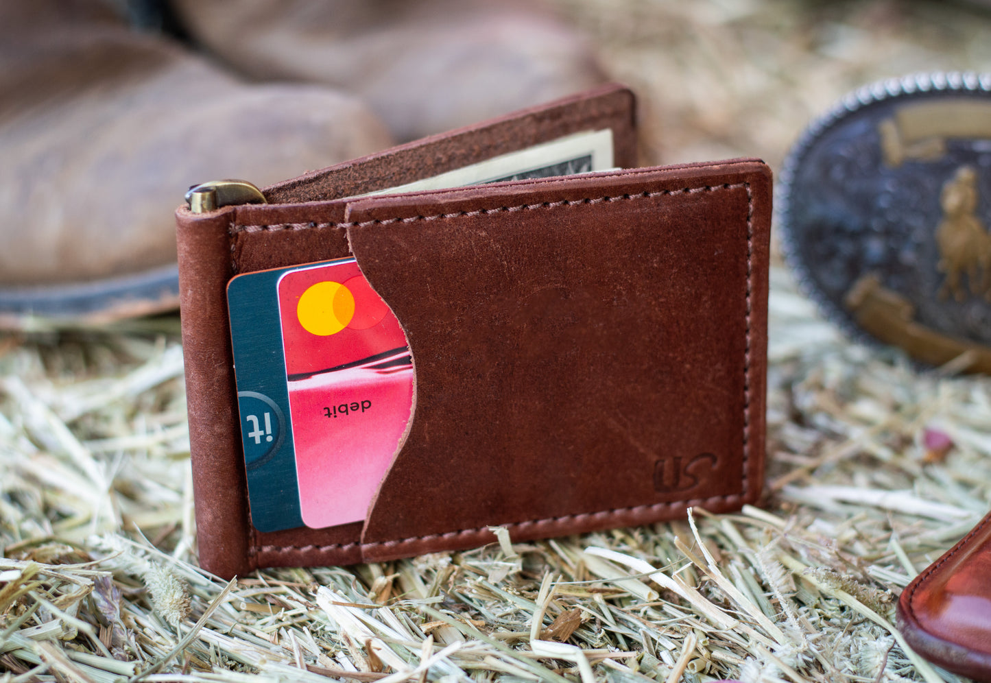 A brown leather wallet sits on top of a hay bale. The wallet shows two credit cards within its exterior card pocket. 