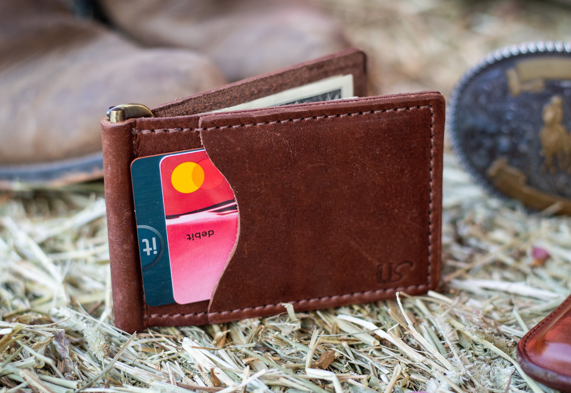 A brown leather wallet sits on top of a hay bale. The wallet shows two credit cards within its exterior card pocket. 