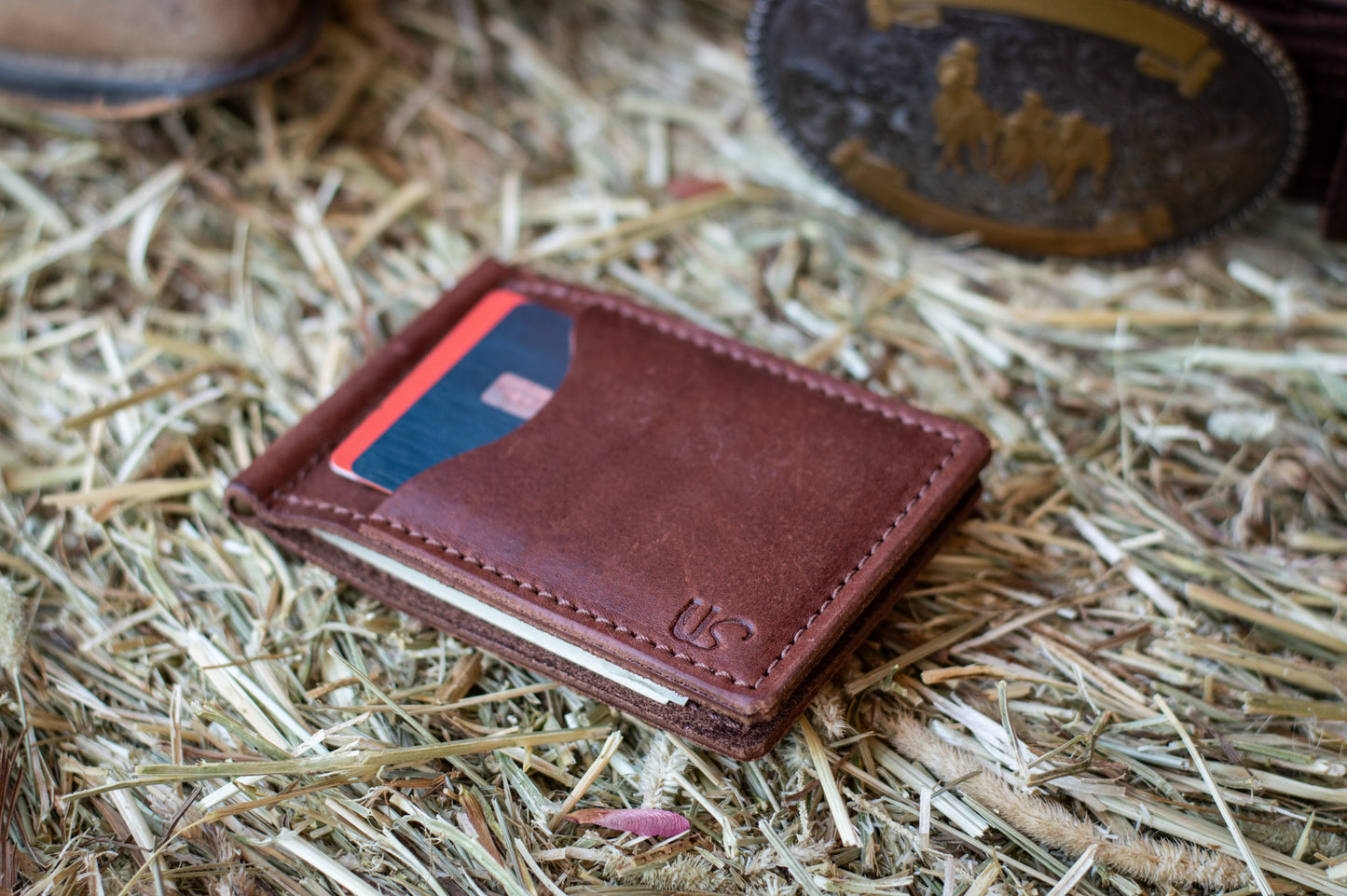 A brown leather bifold money clip wallet lies on a bale of hay. Two credit cards peak out of the exterior card slot. 