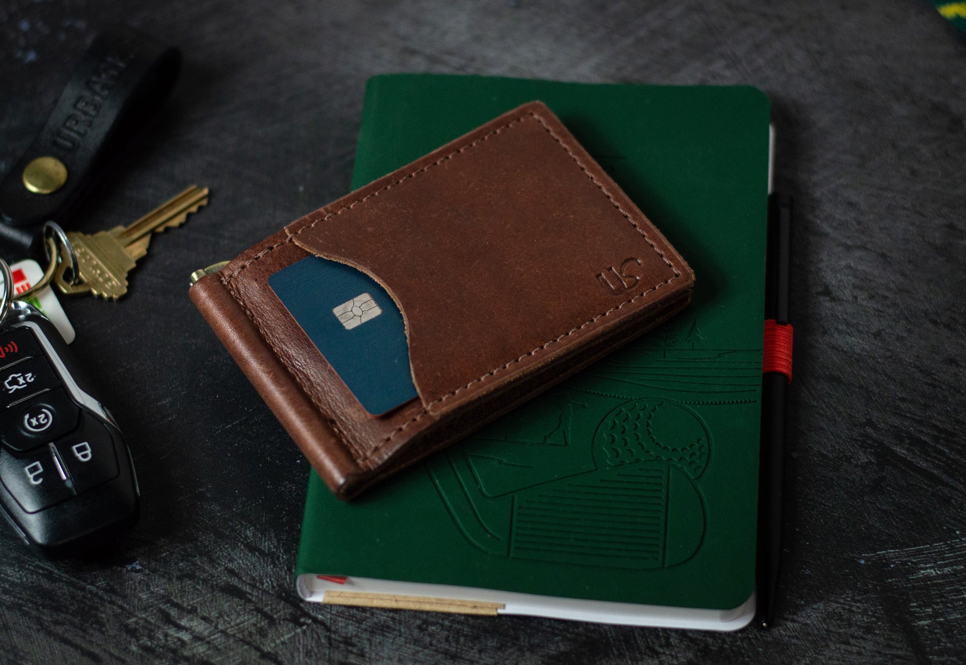 Brown leather wallet, with a credit card in the front card pocket, on a green notebook with keys and a car keychain in the background.