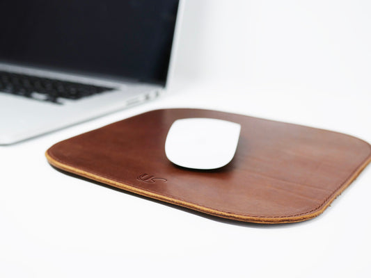 A computer mouse sits on top of a full-grain leather mousepad, while an open laptop sitting on the table is waiting to be powered up.