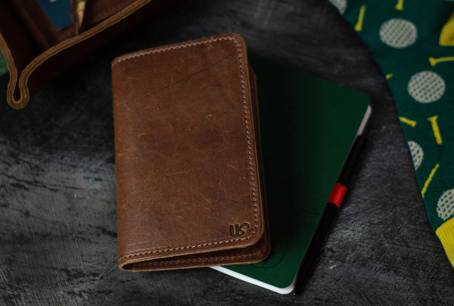 Brown leather notebook on a dark textured surface with a green notebook and colorful fabric.