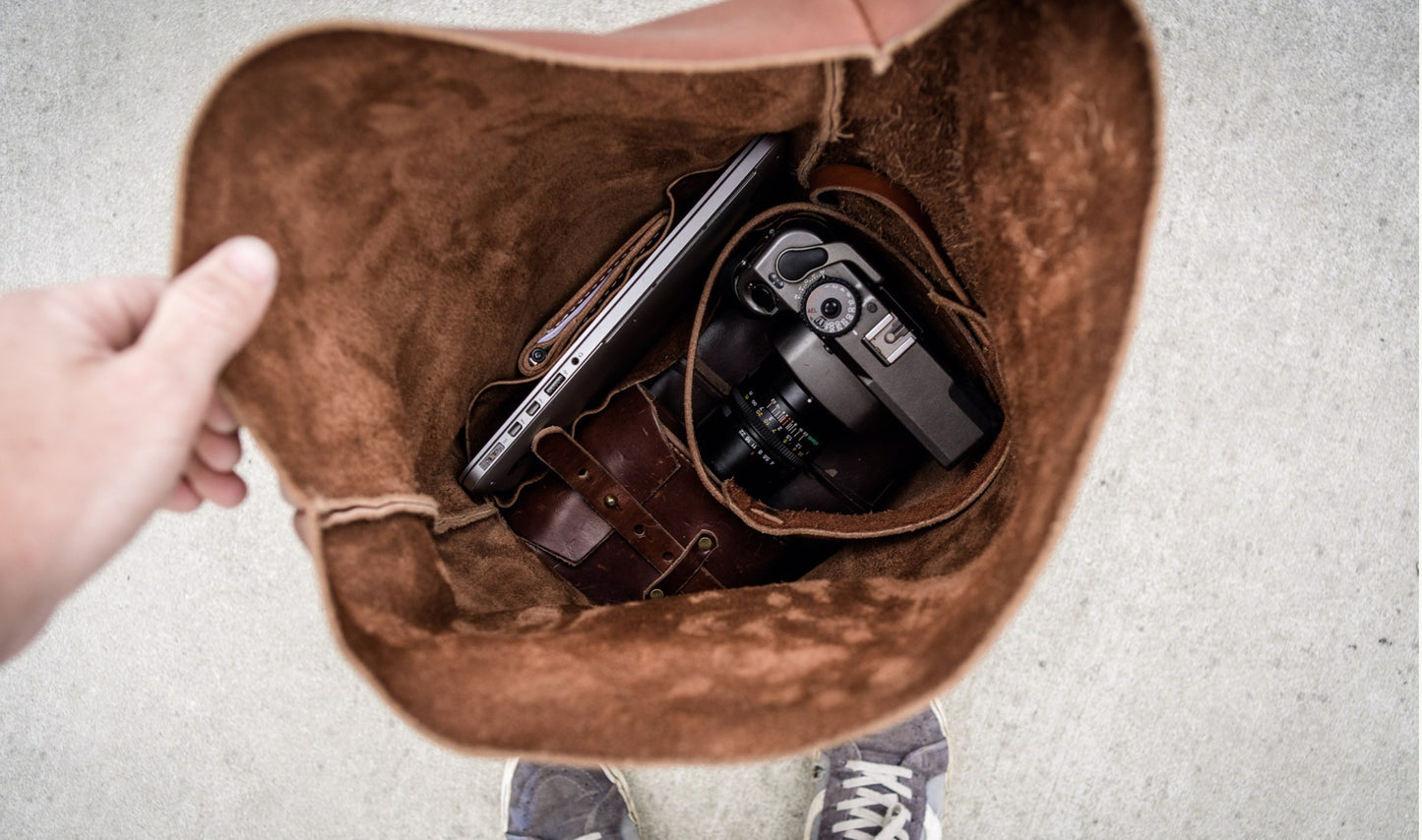 The interior of an open brown leather bag with a phone and camera inside, held by a hand on a light gray background