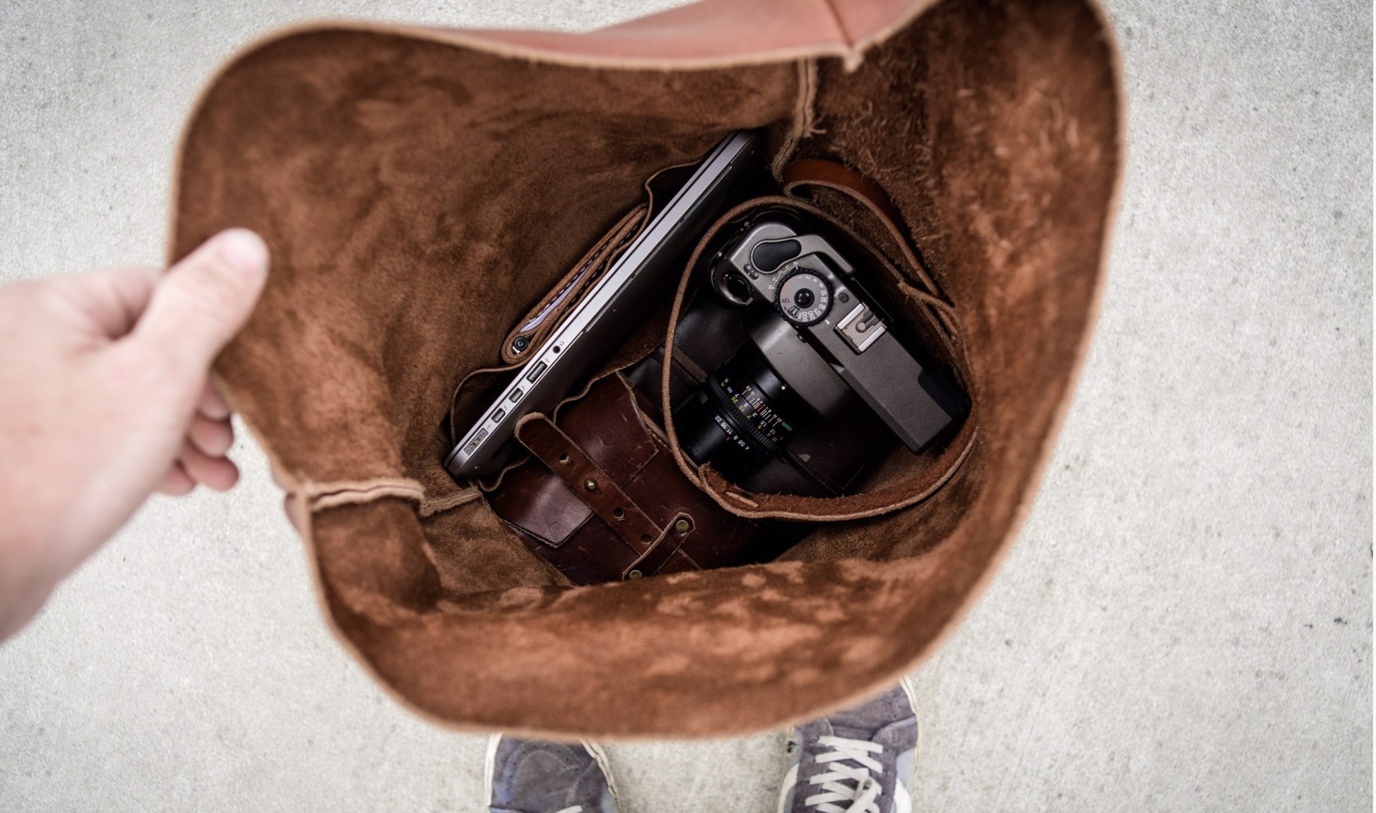 The interior of an open brown leather bag with a phone and camera inside, held by a hand on a light gray background