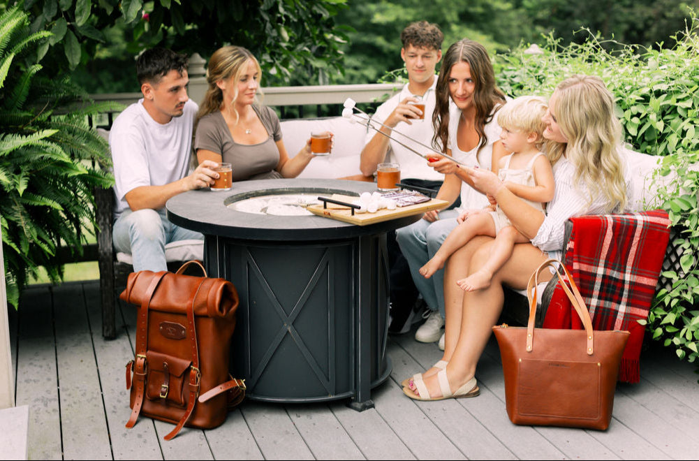 Family gathered around a fire pit on a patio holding leather wrapped glasses, a mens leather backpack & womens leather handbag sits on the ground next to the fire pit.