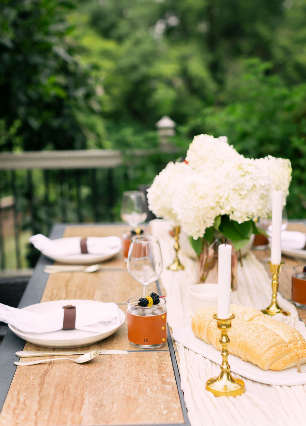 Elegant outdoor dining table setting with white flowers, gold candlesticks, and leather wrapped whiskey glasses