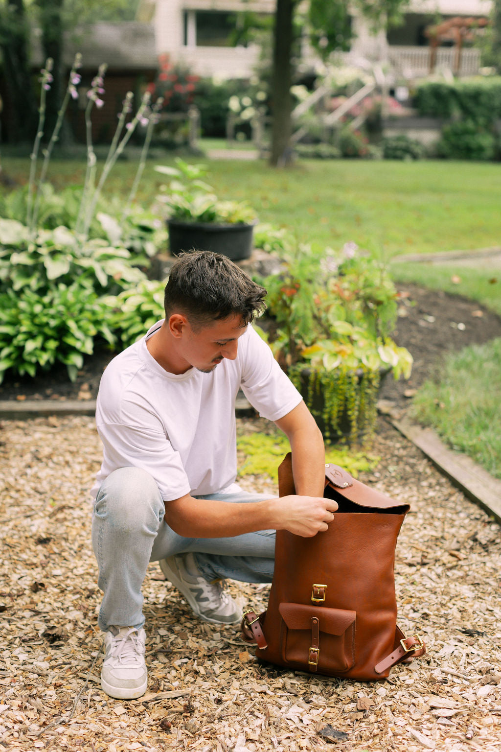 man kneeling next to a brown leather backpack, reaching inside 