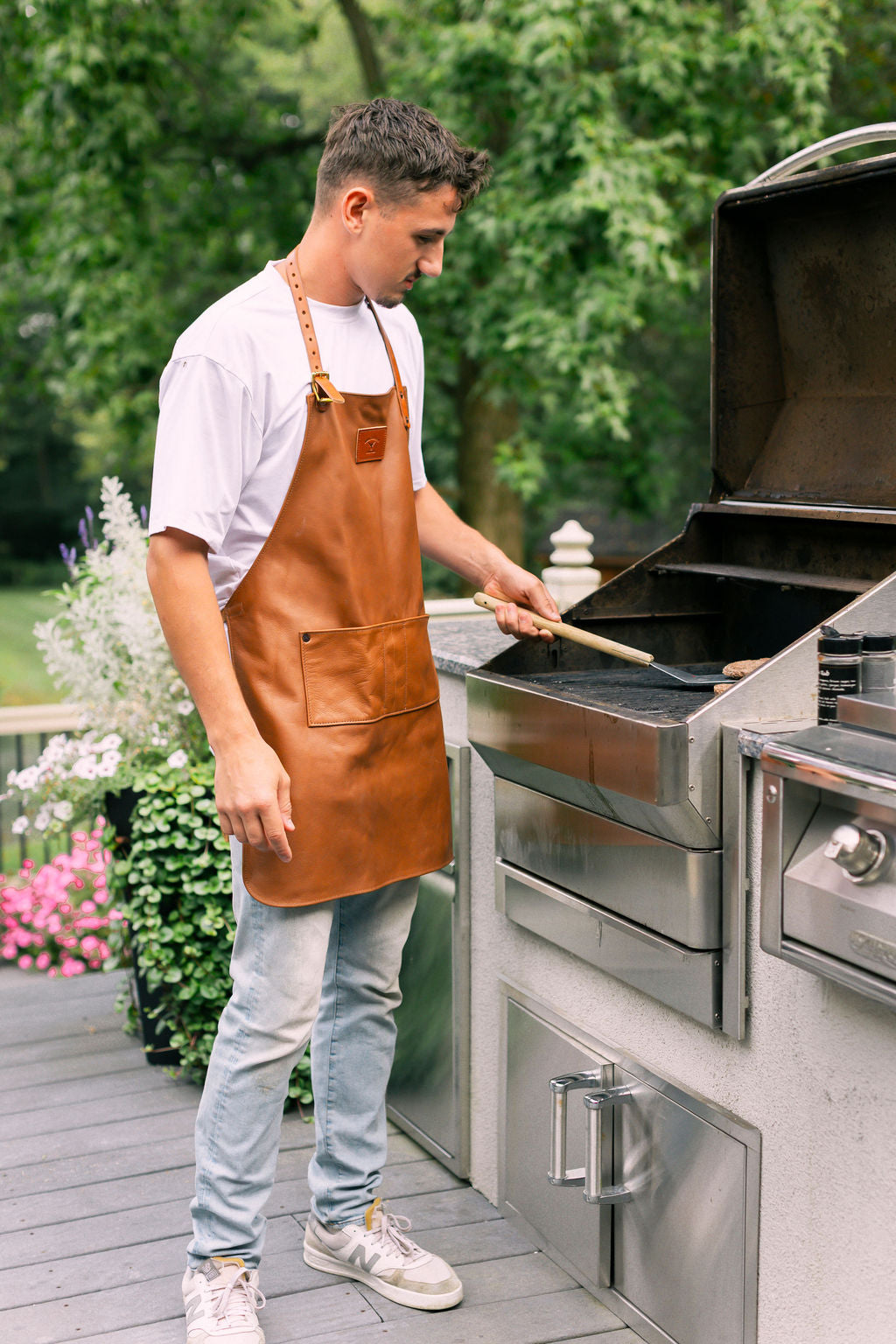 Man grilling outdoors using a stainless steel barbecue grill, wearing a brown leather apron