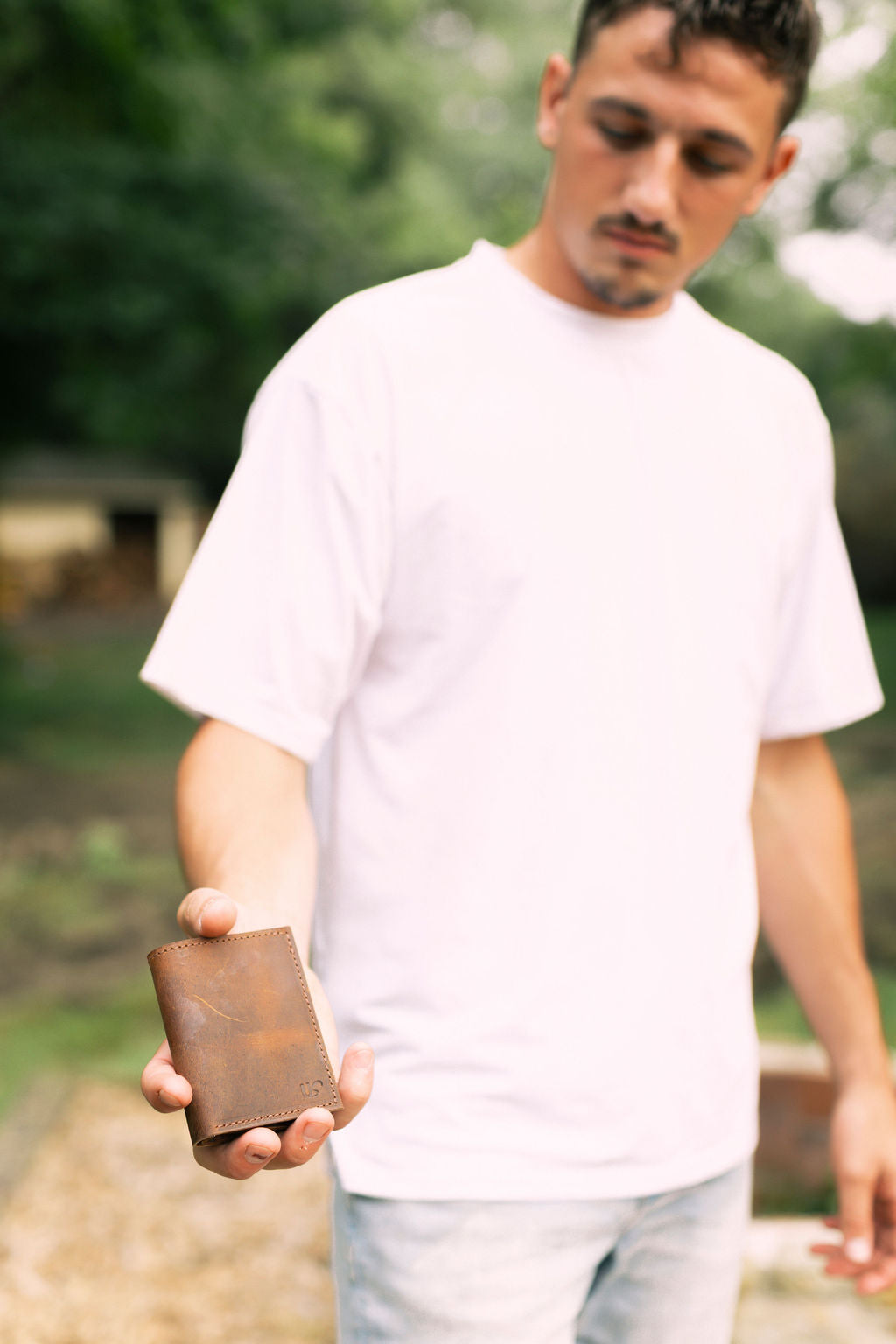 Man holding a brown leather wallet outdoors with greenery in the background