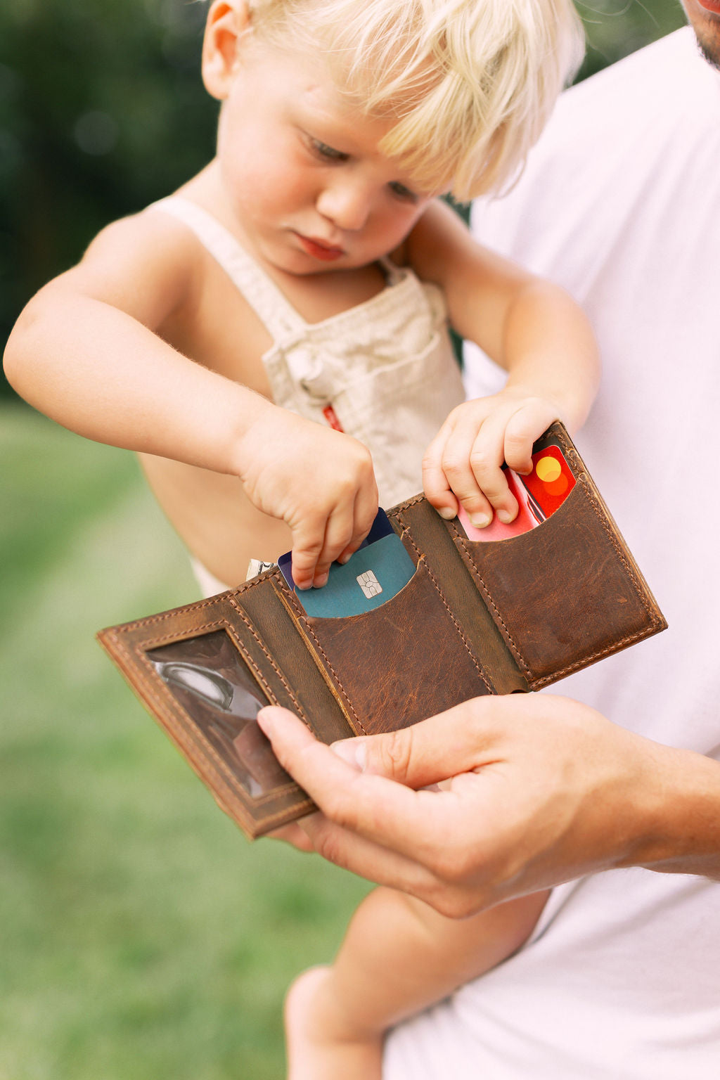 A man holds a small child. The child reaches into an open leather trifold wallet and pulls out a credit card. 
